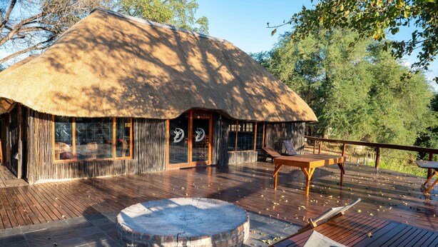 Een gebouw met rieten dak, een houten terras, een vuurkorf en buitenzitplaatsen.