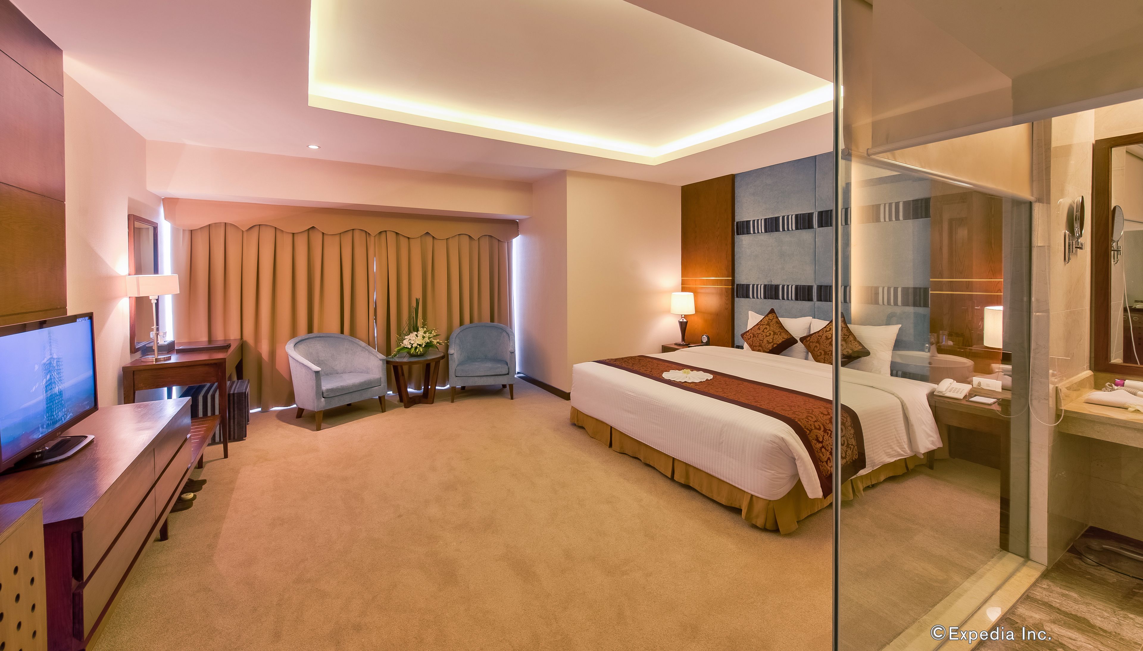 A hotel room with a bed, bedside table, lamp, chair, TV, and a glass-enclosed bathroom.