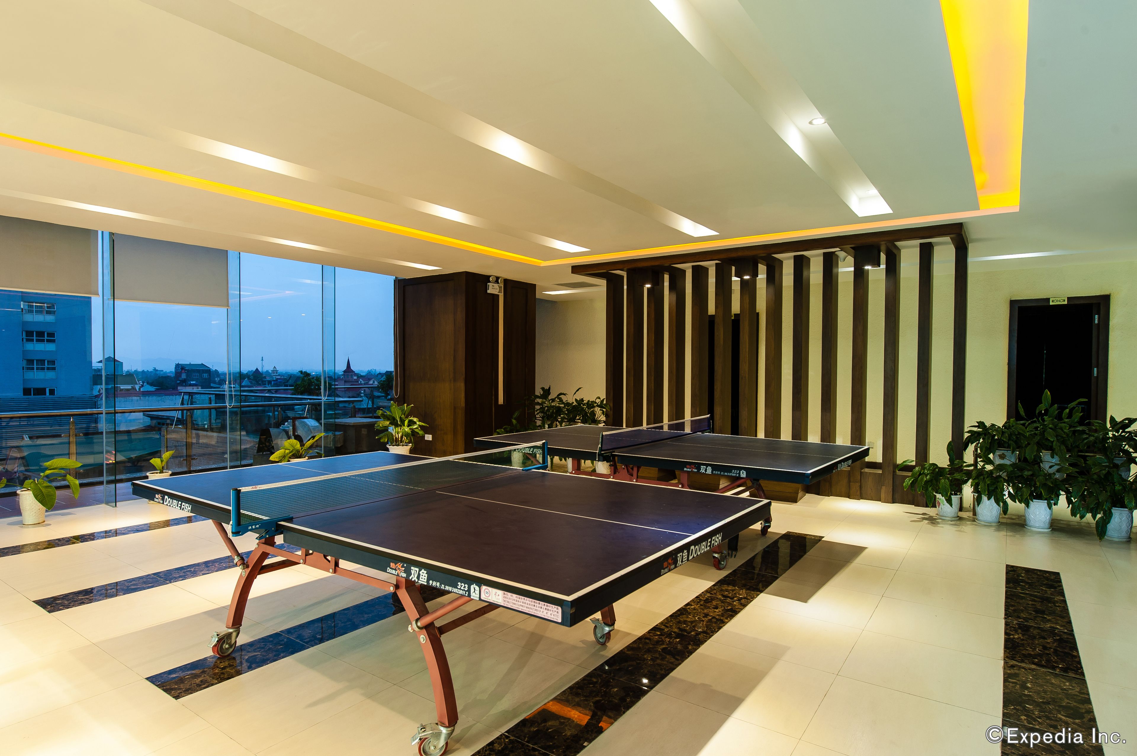 A modern indoor ping pong table area with multiple tables, potted plants, and a city view through large windows.