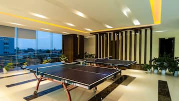A modern indoor ping pong table area with multiple tables, potted plants, and a city view through large windows.