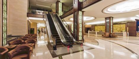 A spacious lobby with a central escalator, seating area with leather chairs, and decorative lighting.