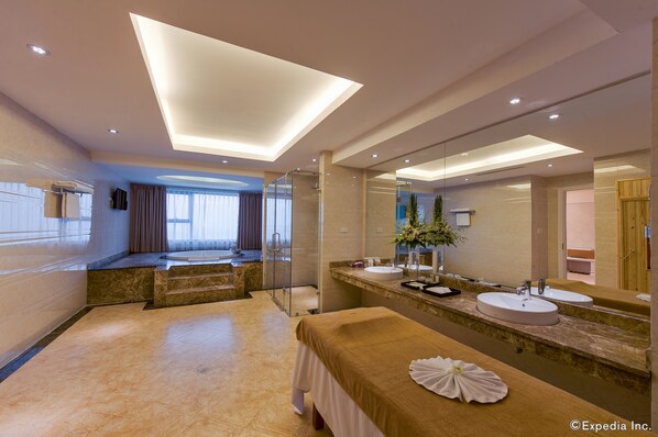 Un baño de hotel moderno con un espejo grande, dos lavamanos, una camilla de masaje y una zona de ducha.