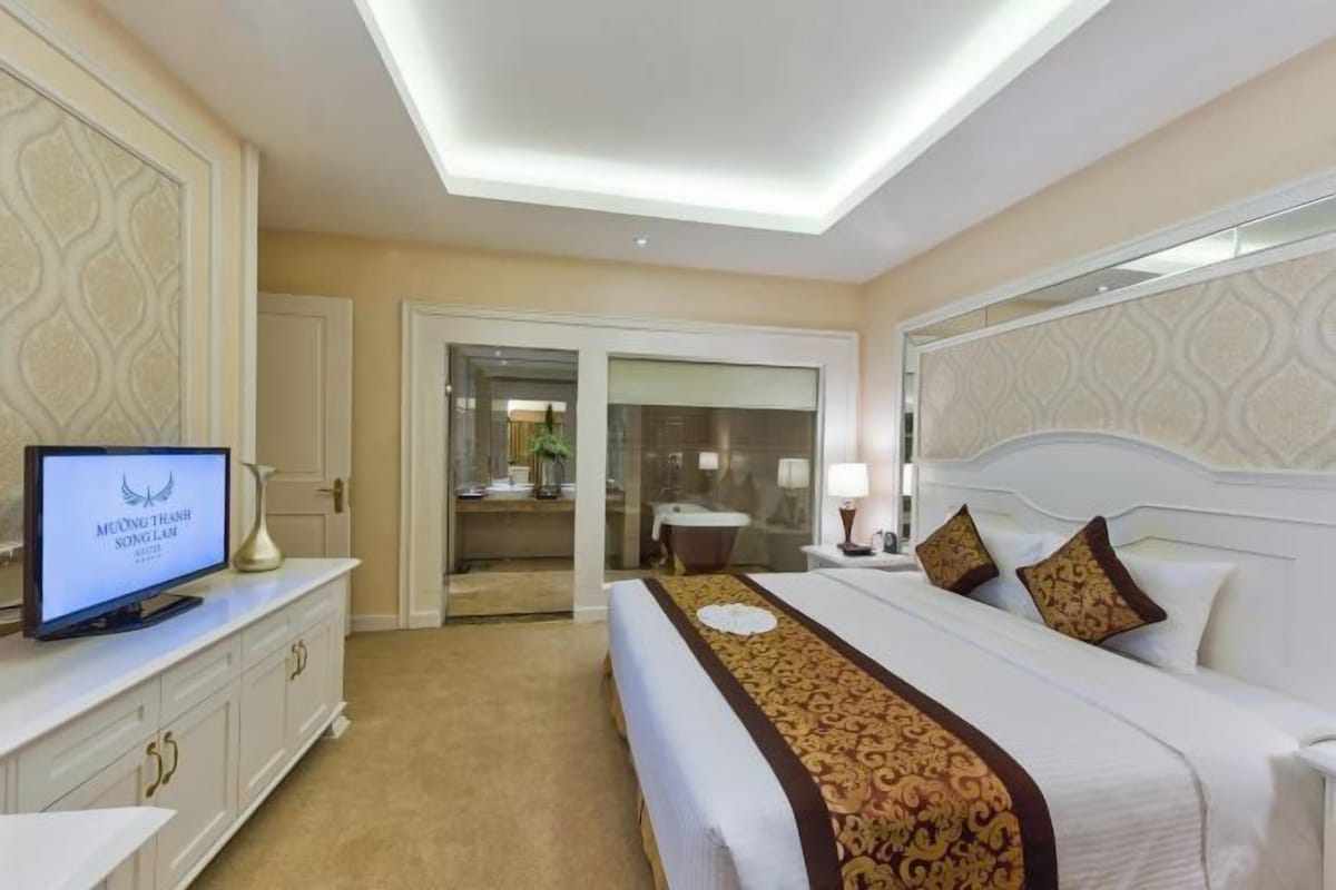Habitación de hotel con cama, televisor, escritorio y baño visible a través de una puerta abierta.