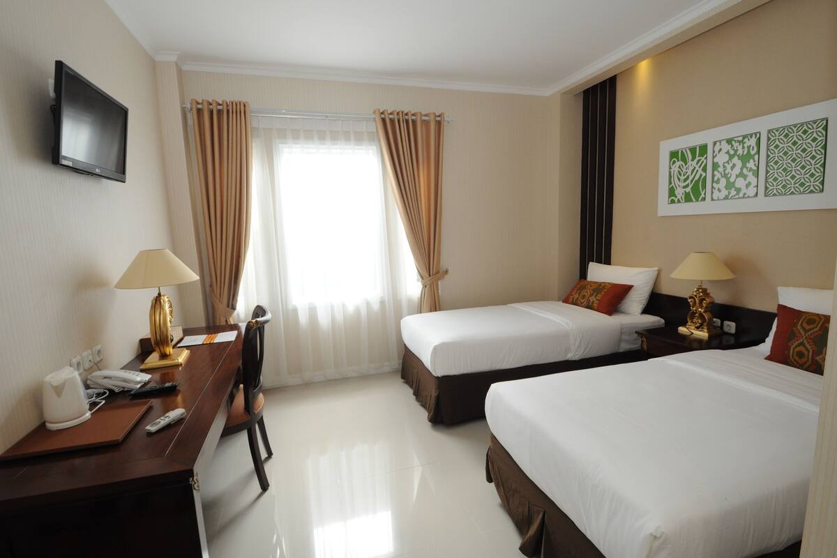 A hotel room with two single beds, a desk with a lamp, a TV, and a window with curtains.