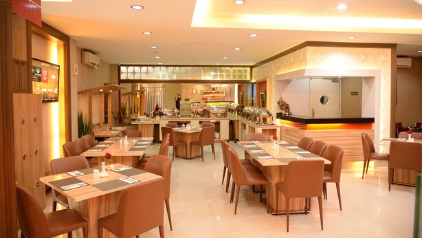 A modern restaurant with multiple dining tables and chairs, a kitchen area, and a reception desk. - Rangkayo Basa Halal Hotel (Padang)