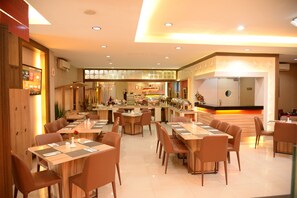 A modern restaurant with multiple dining tables and chairs, a kitchen area, and a reception desk. - Rangkayo Basa Halal Hotel (Padang)
