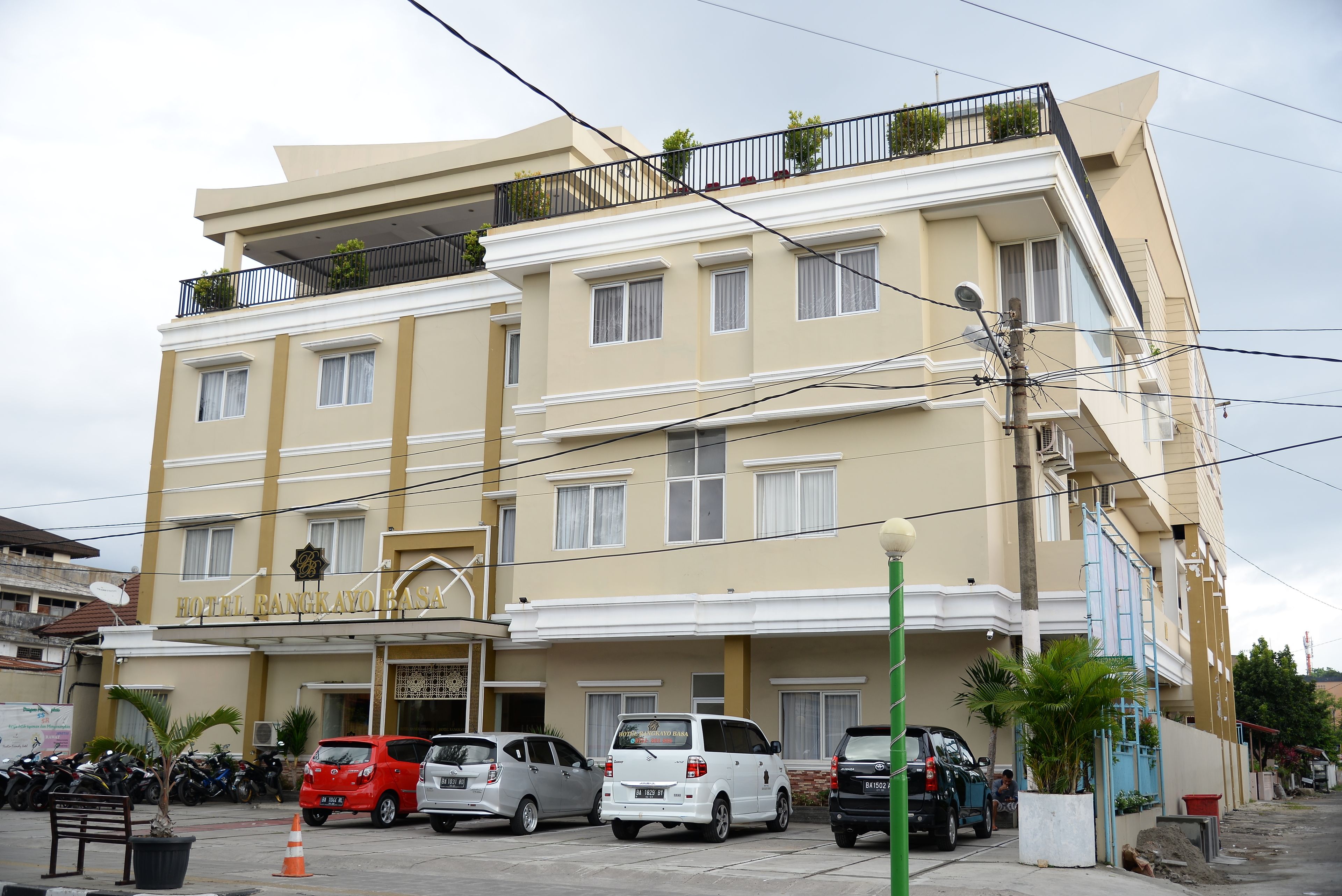 A multi-story hotel with a flat facade, balconies with plants, and a parking area with several cars.