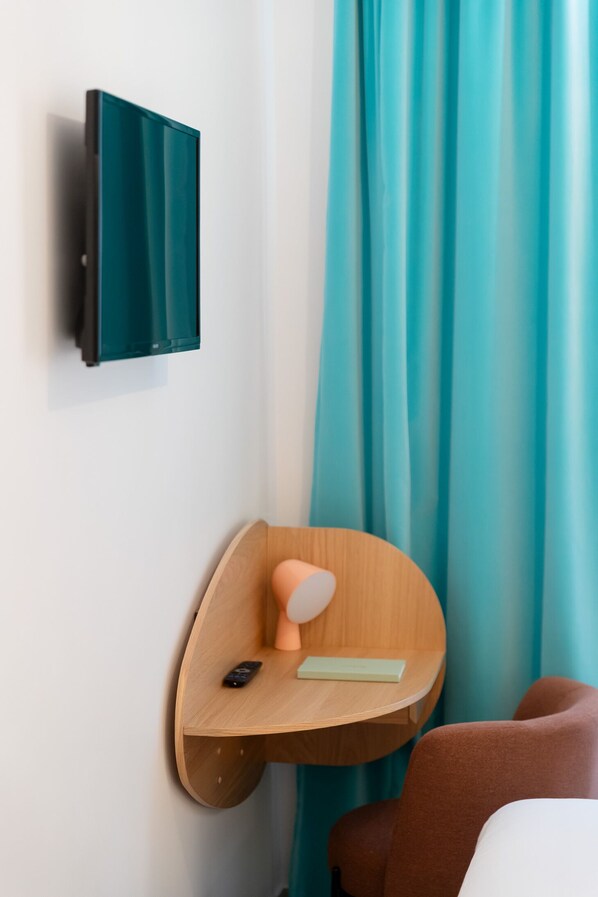 Une chambre moderne avec un rideau turquoise, un téléviseur fixé au mur, une table d’appoint en bois avec une télécommande, et un fauteuil marron.