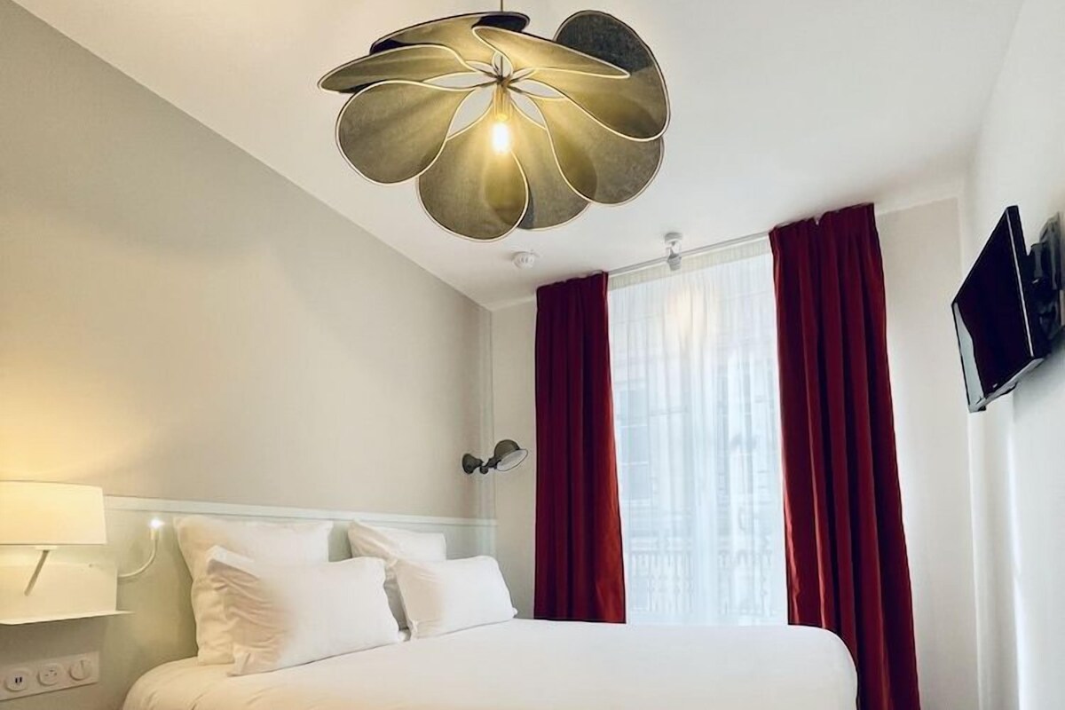 A hotel room with a large bed, a modern chandelier, a flat-screen TV, and red curtains.