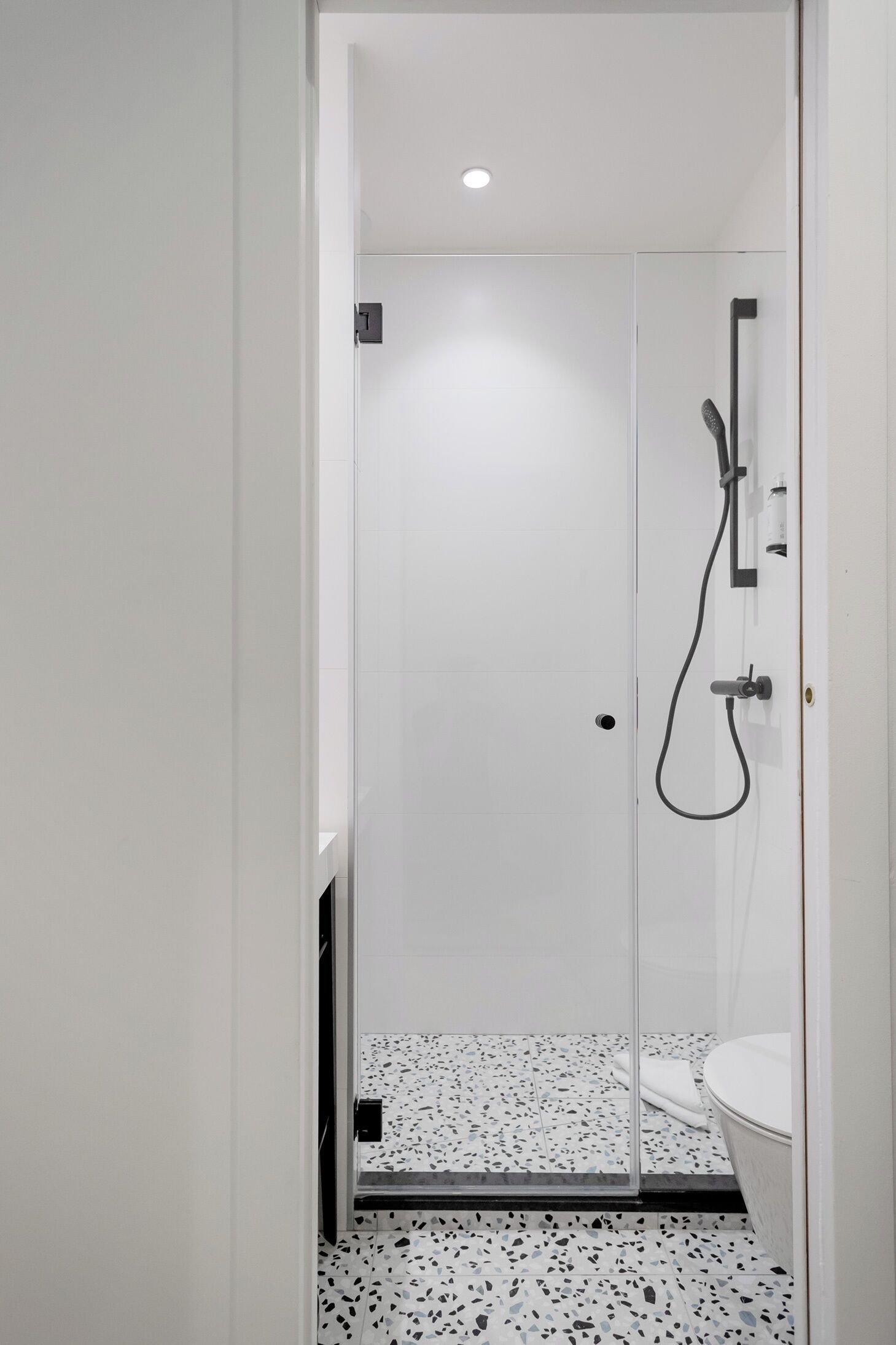 Een smalle badkamer met een douchecabine van glas, een wit toilet en een geruite vloer.