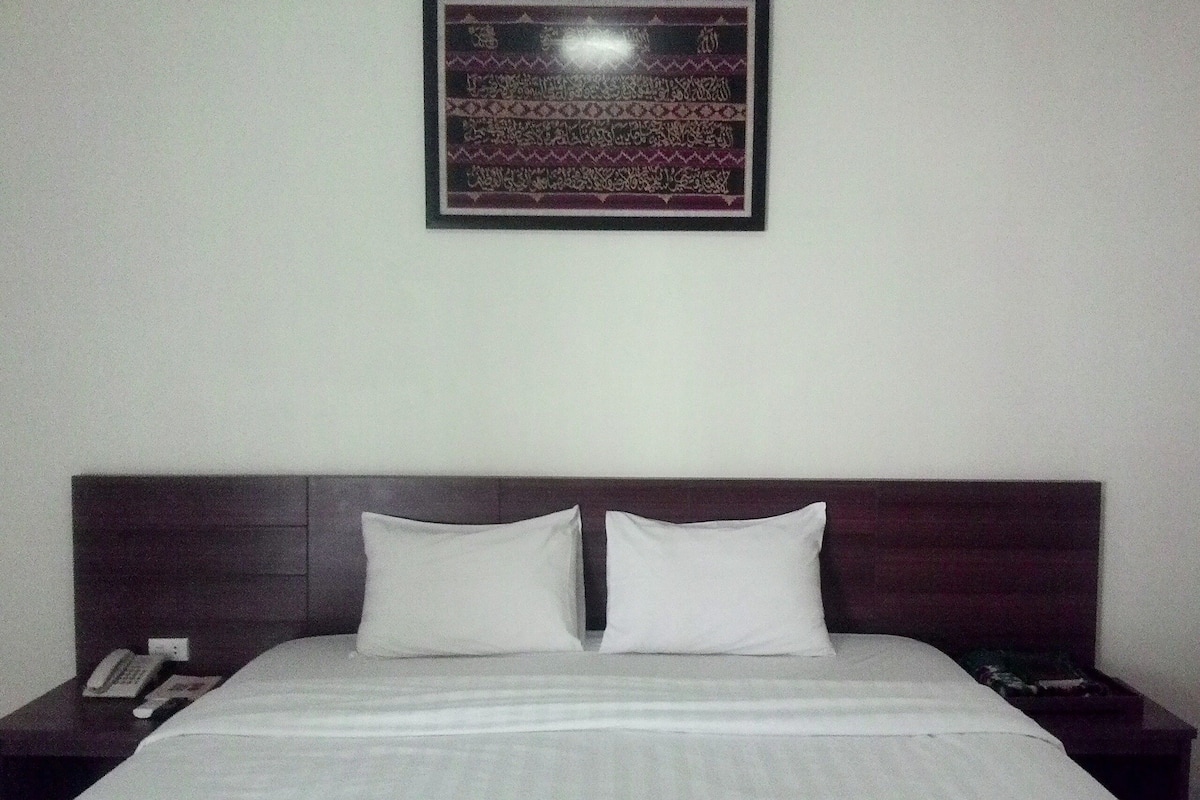 A hotel room with a bed, two pillows, a nightstand with a phone, and a framed artwork on the wall.