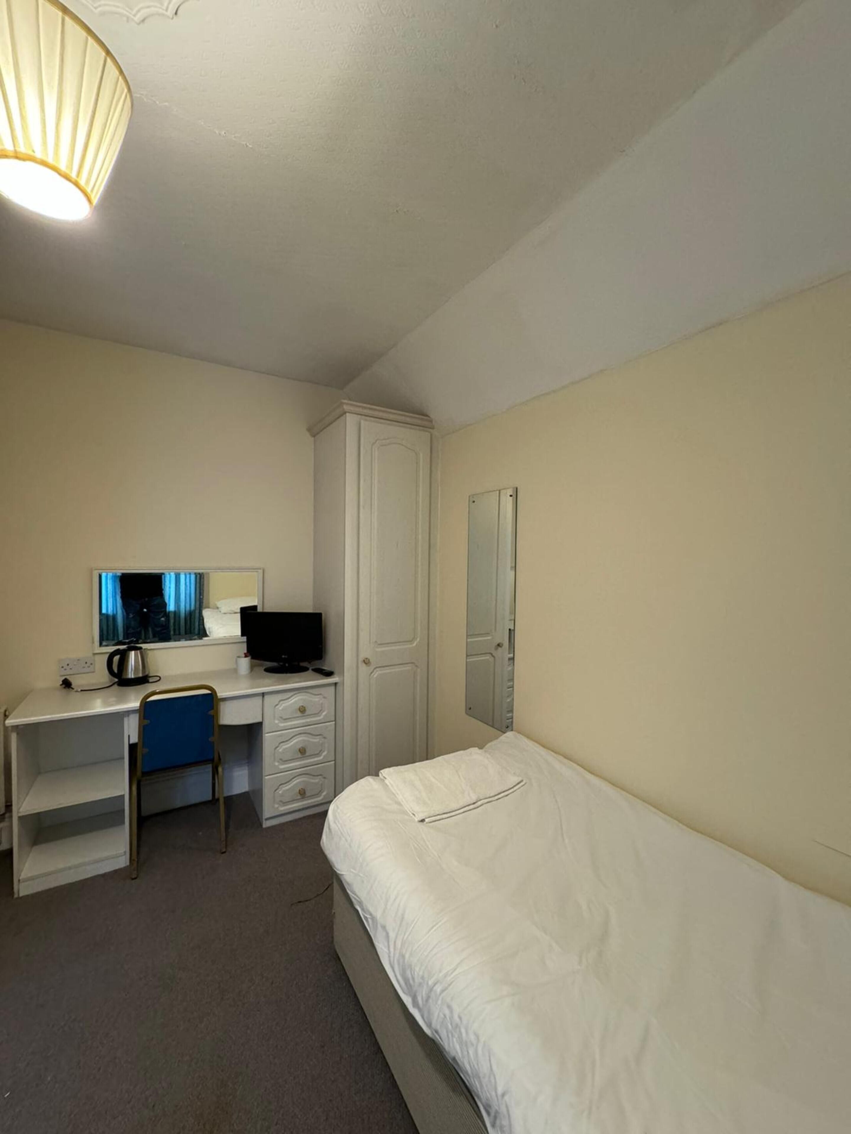 A compact hotel room with a single bed, a desk with a computer, a chair, a wardrobe, and a mirror.