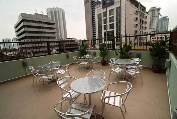 Una terraza en la azotea con mesas y sillas de metal, plantas en macetas y vistas de edificios de la ciudad.