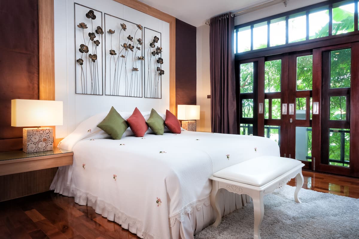 Een groot bed met een witte spreideken en meerdere kussens, een nachtkastje met een lamp, en uitzicht op groen door grote ramen.