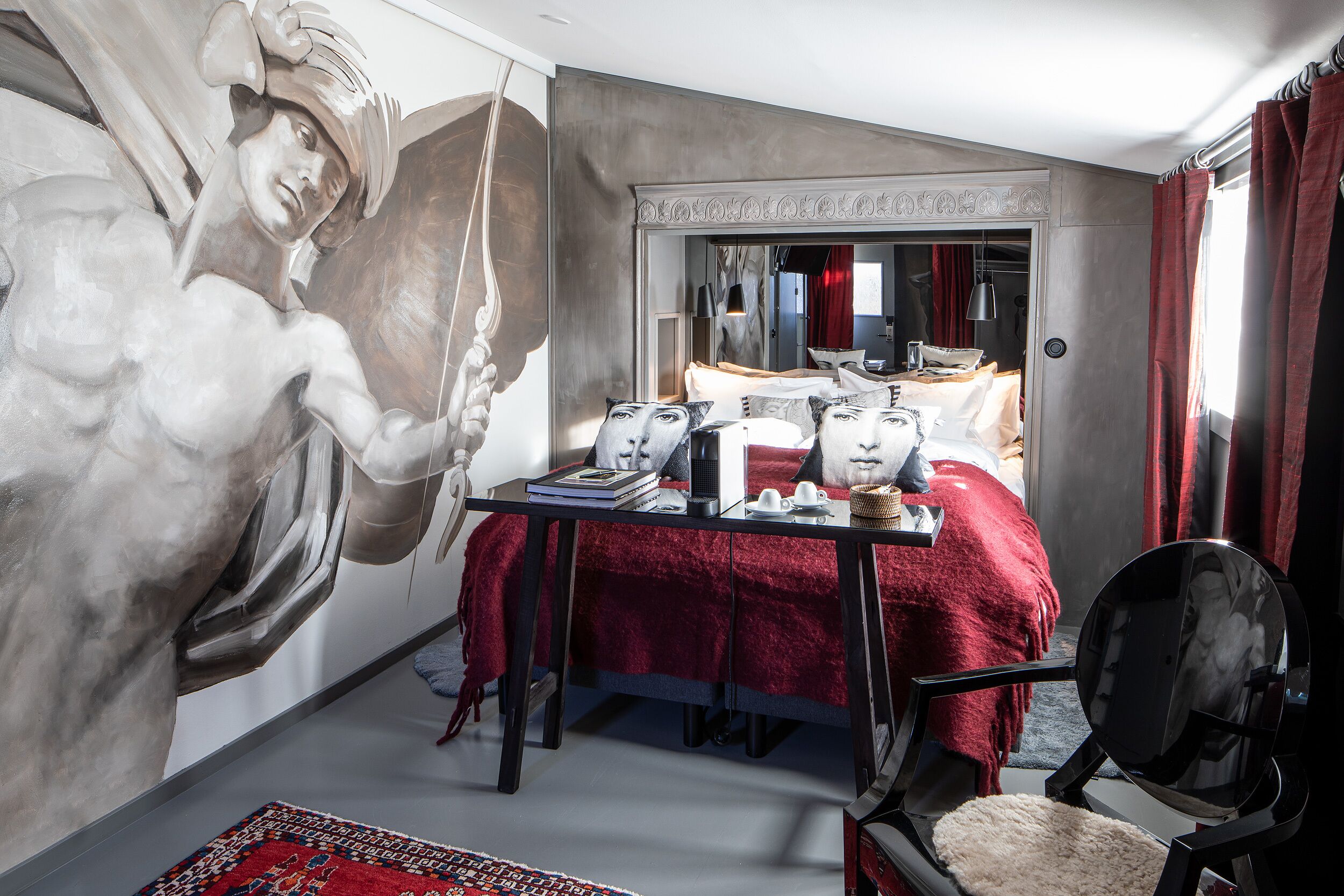 A bedroom with a large mural, a bed with red bedding, a desk with books and a lamp, and a chair with a fluffy seat.