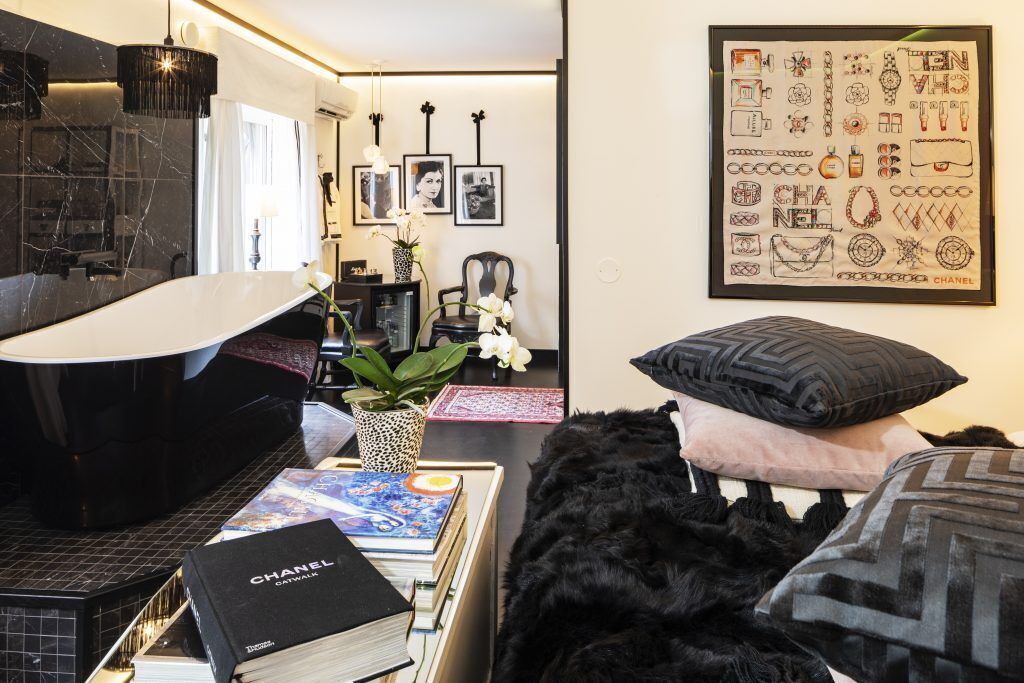 Un dormitorio moderno con una bañera negra, una cama con almohadas negras y rosas, un cartel enmarcado de la colección Chanel y una estantería con libros y un libro de Chanel.
