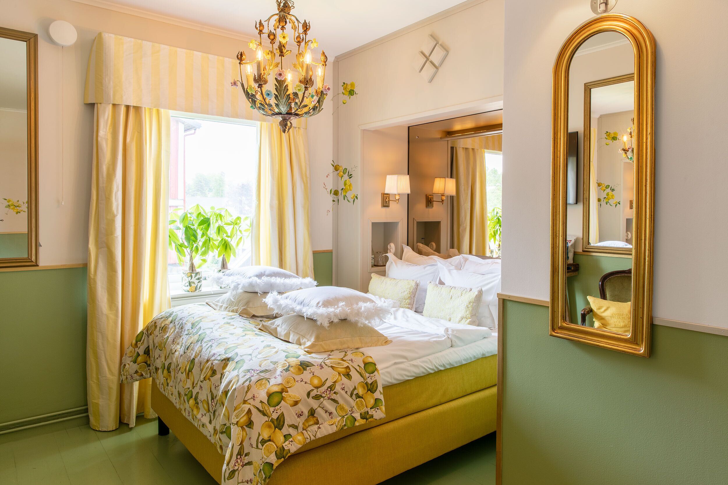 A bedroom with a bed, curtains, a chandelier, a mirror, and a window with plants.