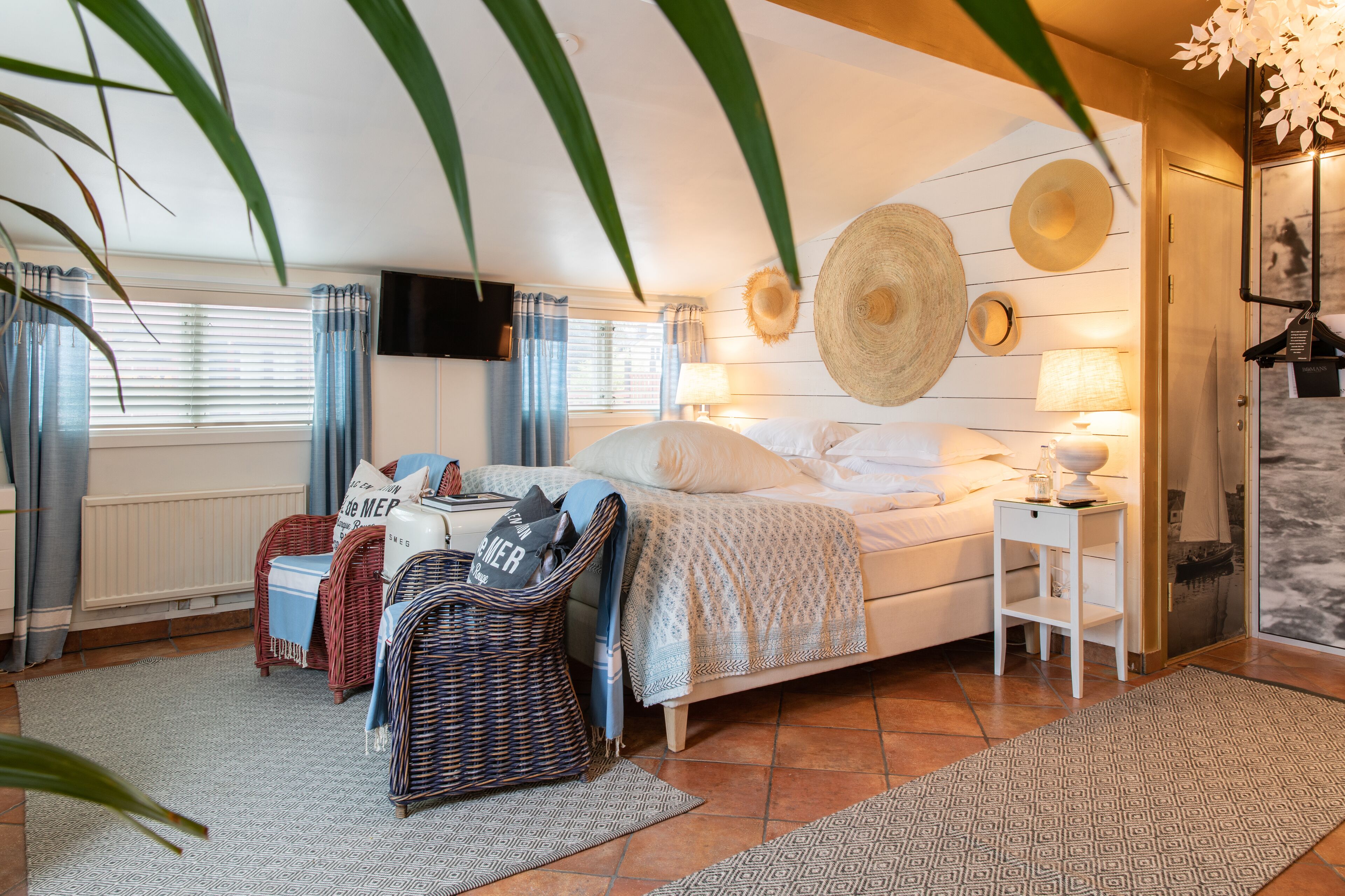 A bedroom with a bed, two nightstands, a wicker chair, a small table, and a wall-mounted TV.