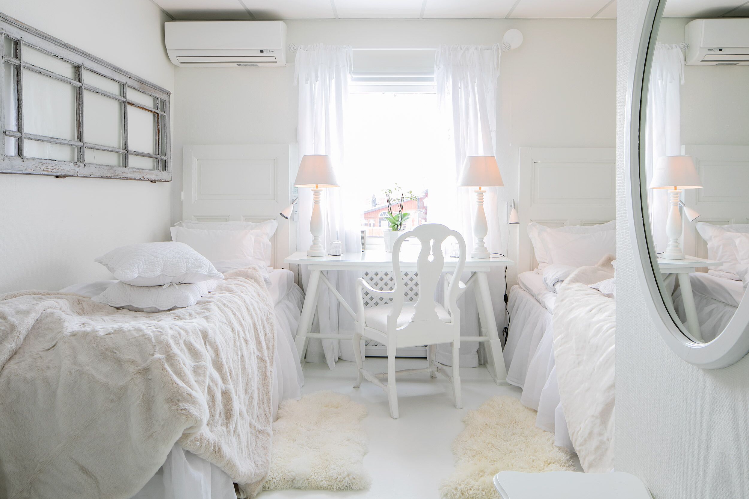 A white bedroom with two beds, a white chair, a small table, and a mirror.