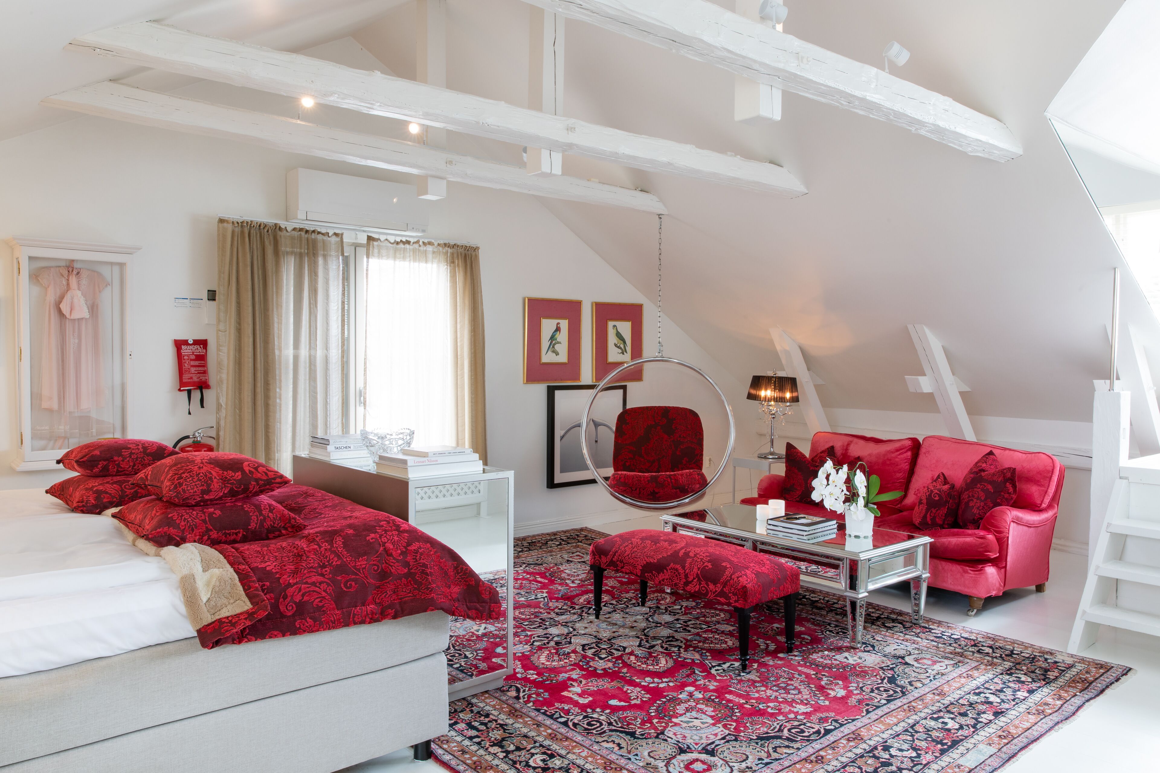 Un dormitorio con una cama grande, un sofá rojo, un sillón redondo y una alfombra estampada.