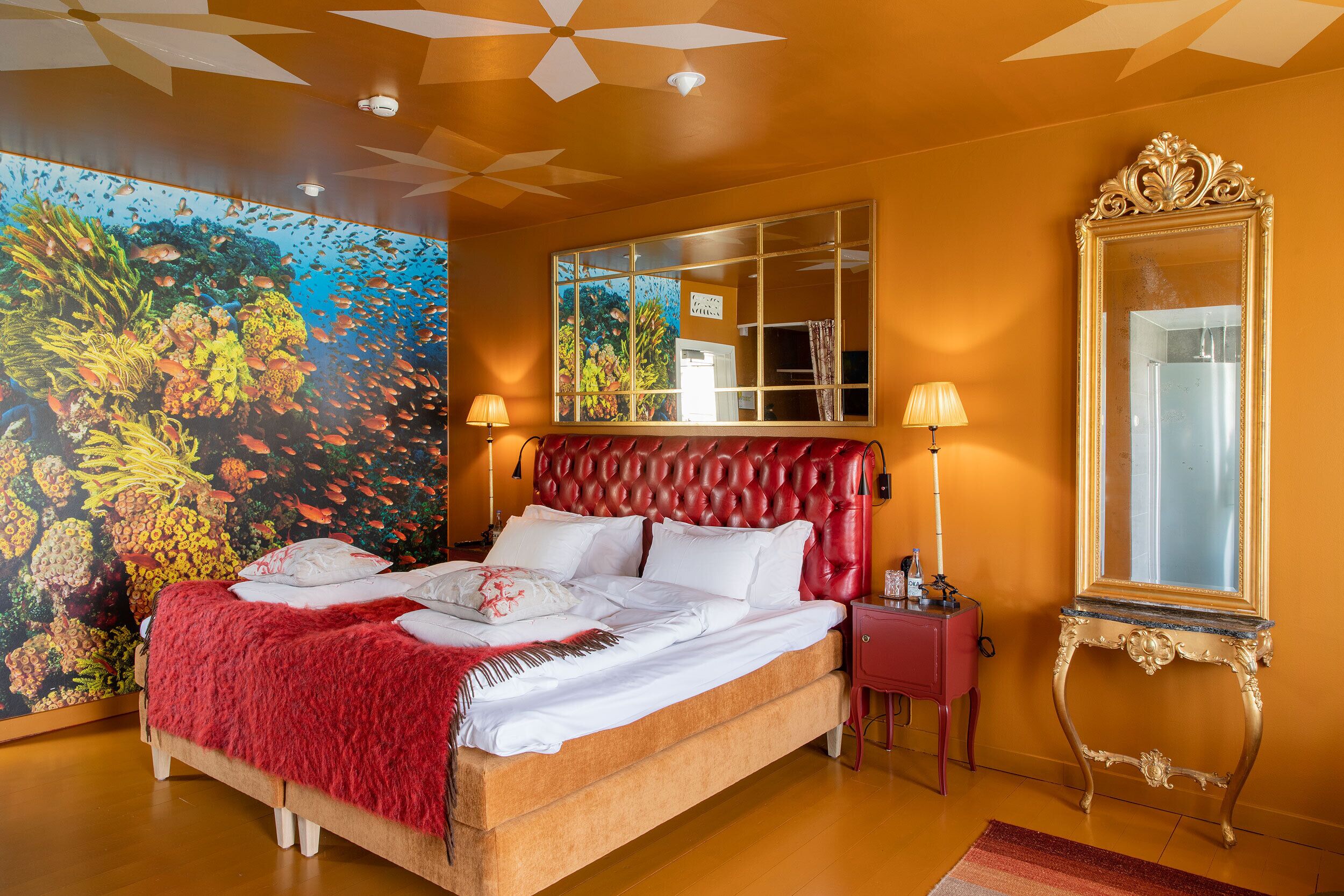 A large bed with a red headboard, a red nightstand, a gold-framed mirror, and a vibrant coral reef mural on the wall.