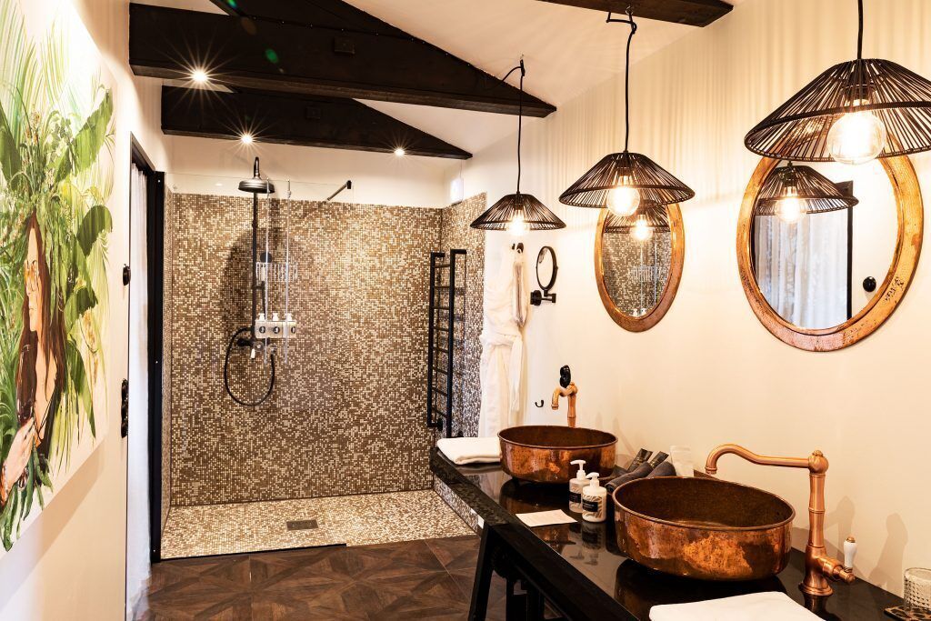 Un baño moderno con un espejo grande, dos lavamanos con detalles de cobre y una zona de ducha con pared texturizada.