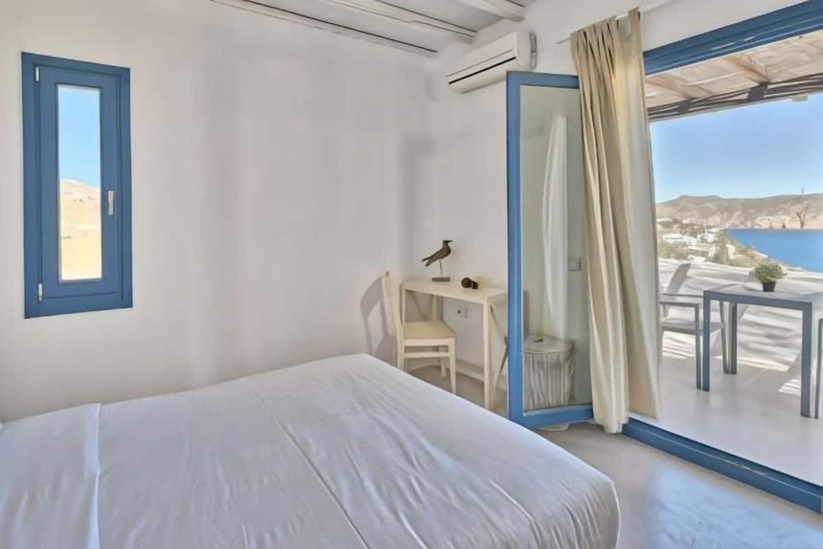 A bedroom with a bed, a desk, a chair, and a balcony with a view of the sea.