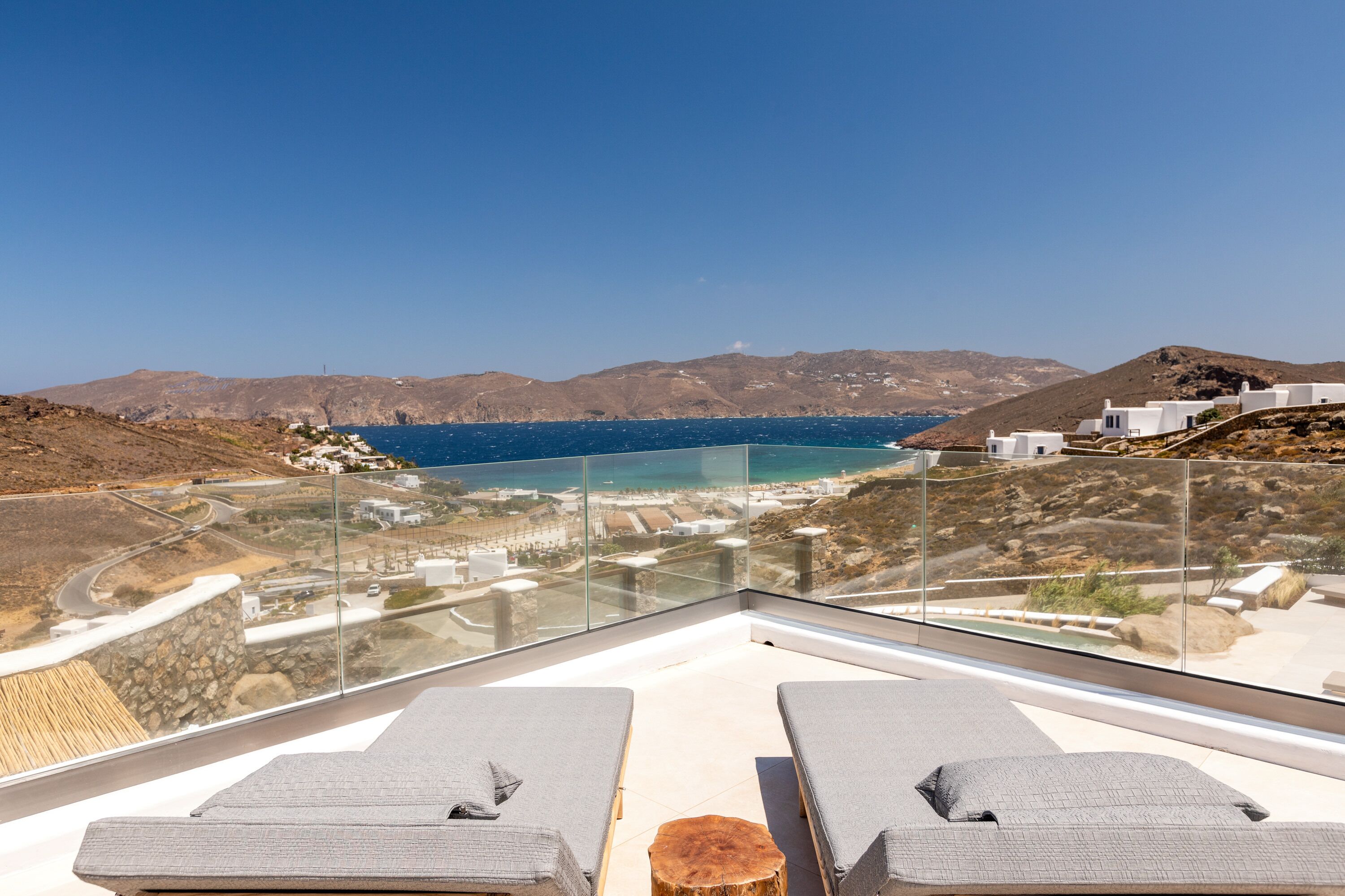 A terrace with two lounge chairs, a glass balustrade, and a view of the sea and distant landscape.