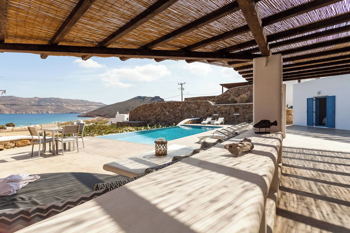 A pool area with lounge chairs, a covered seating area, and a view of the sea and hills.