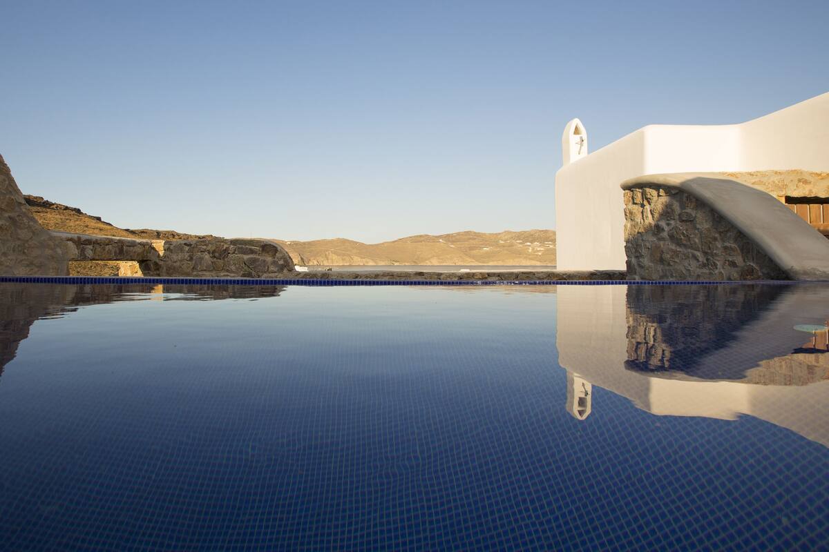 A clear blue infinity pool with a stone wall on one side and a white building with a bell tower on the other.