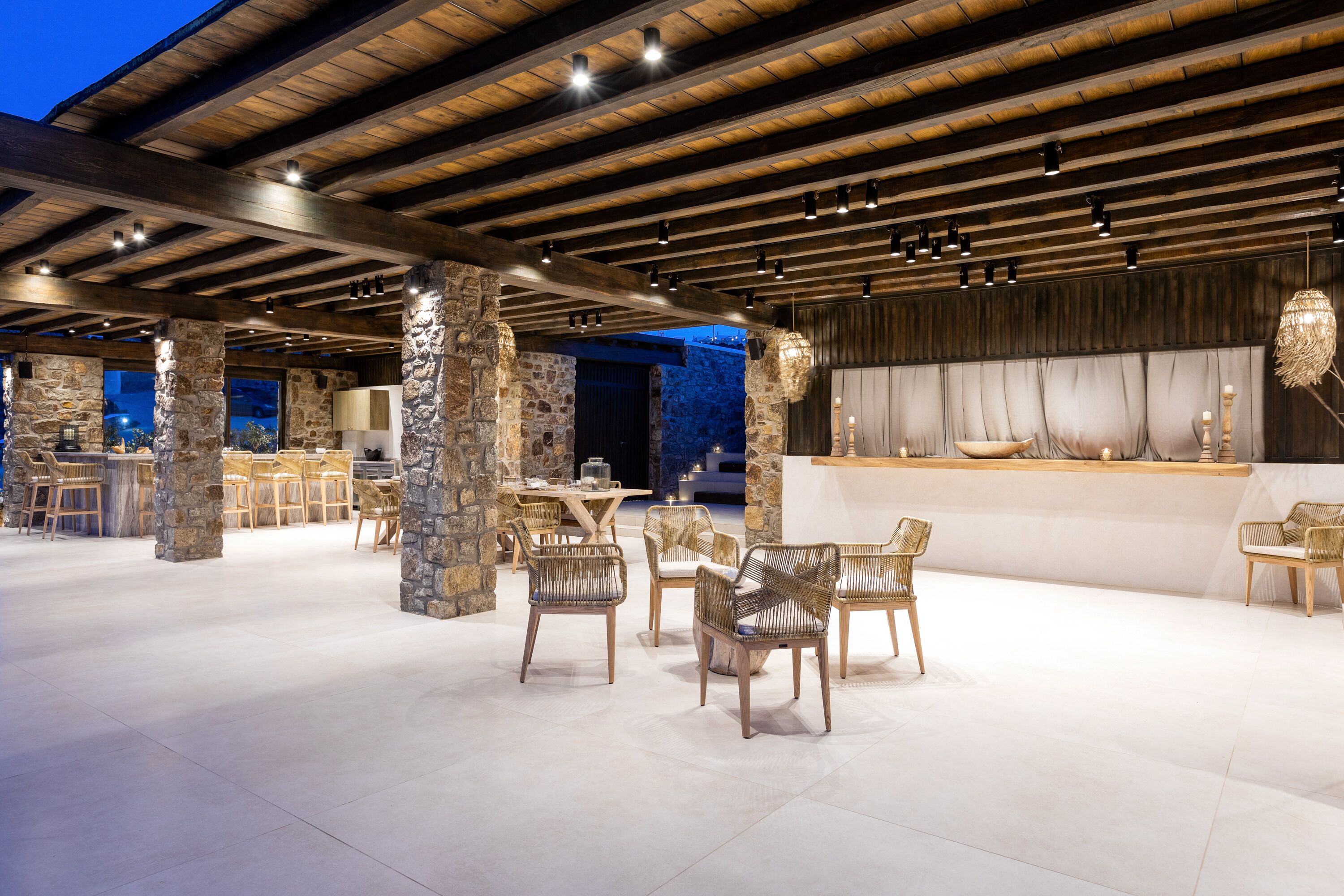 An outdoor dining area with stone pillars, wooden ceiling beams, and wicker chairs.