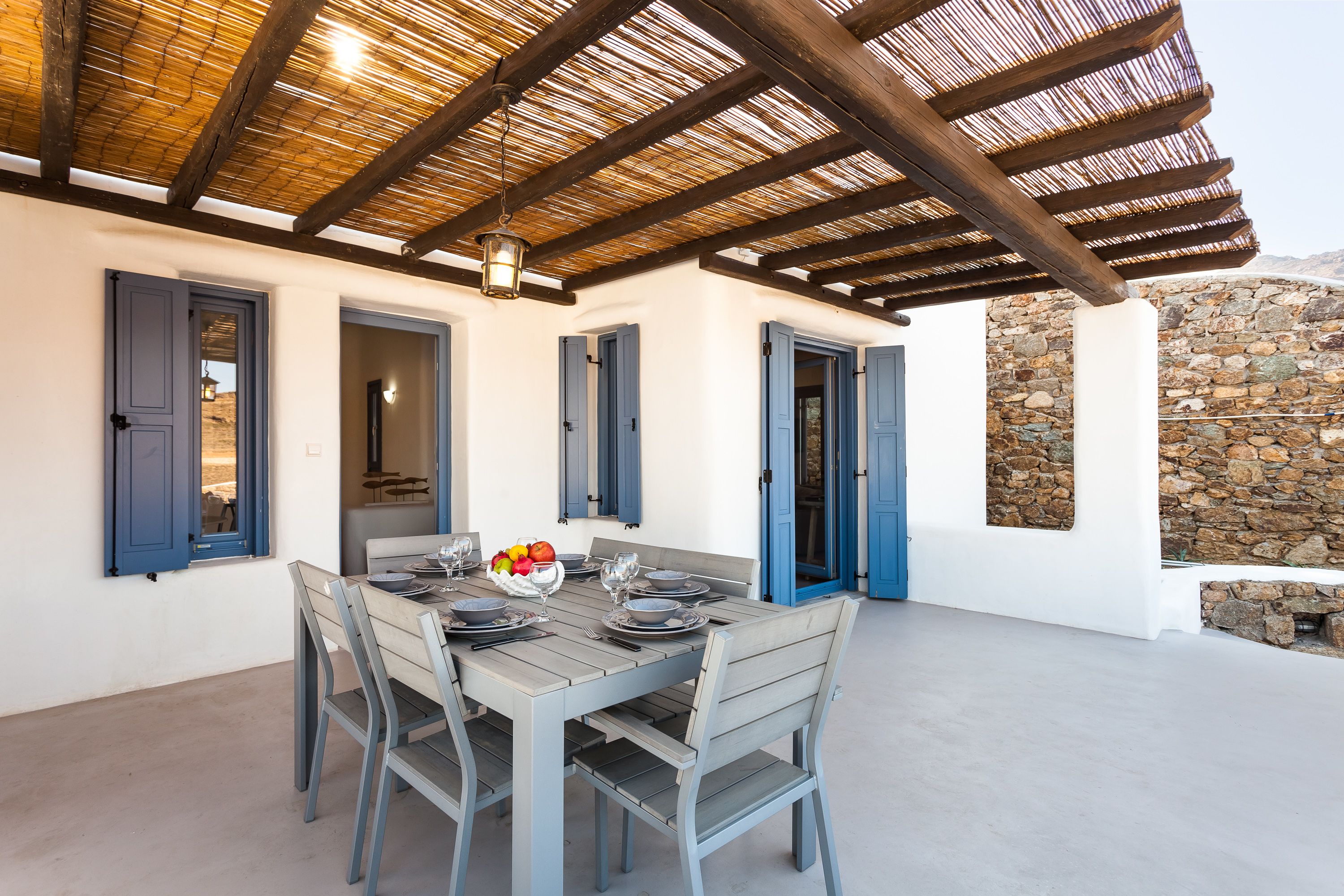 An outdoor dining area with a wooden table set for four, blue shutters, and a stone wall in the background.