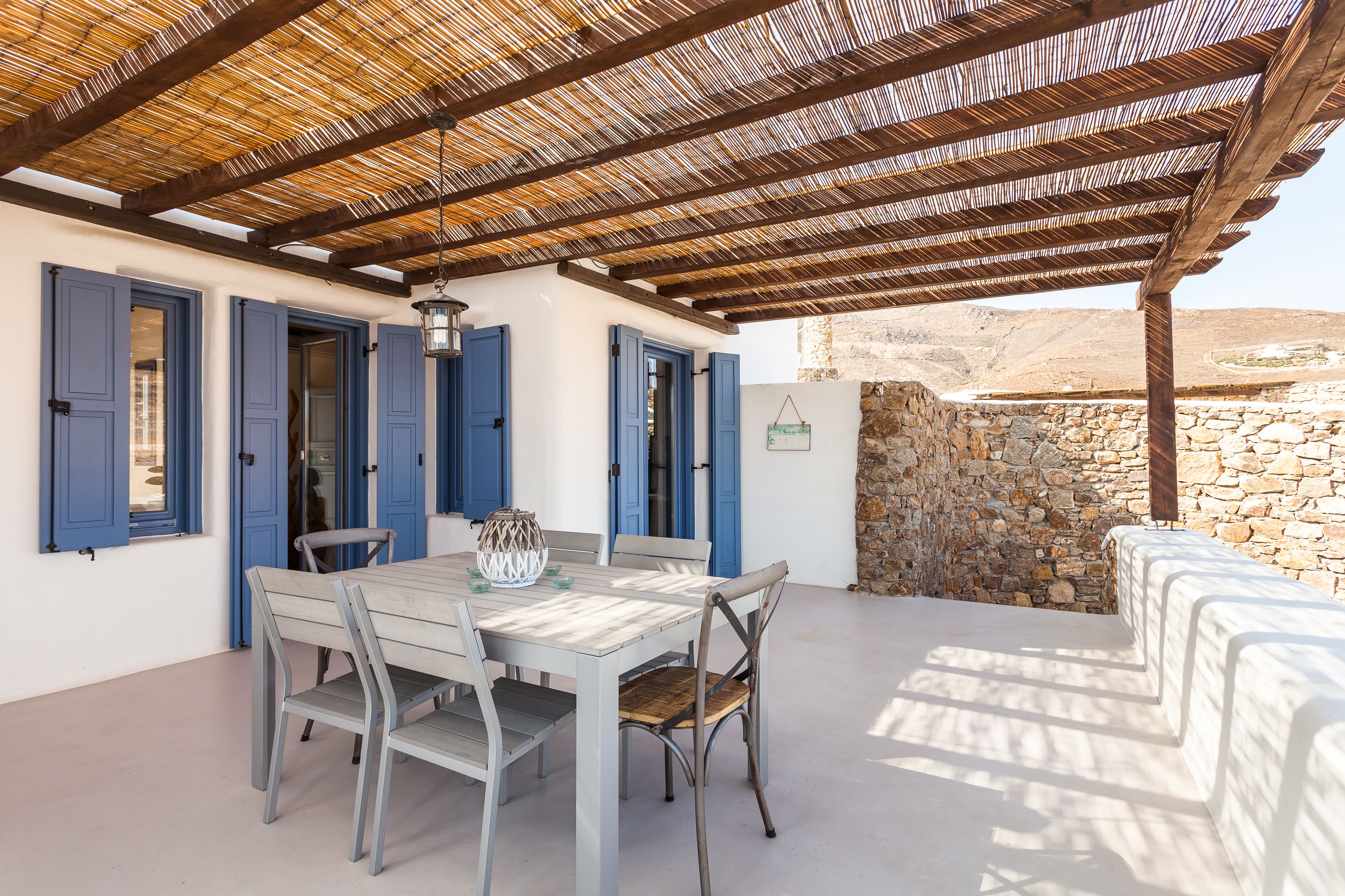 A covered outdoor dining area with a table and chairs, a stone wall, and a view of the landscape.