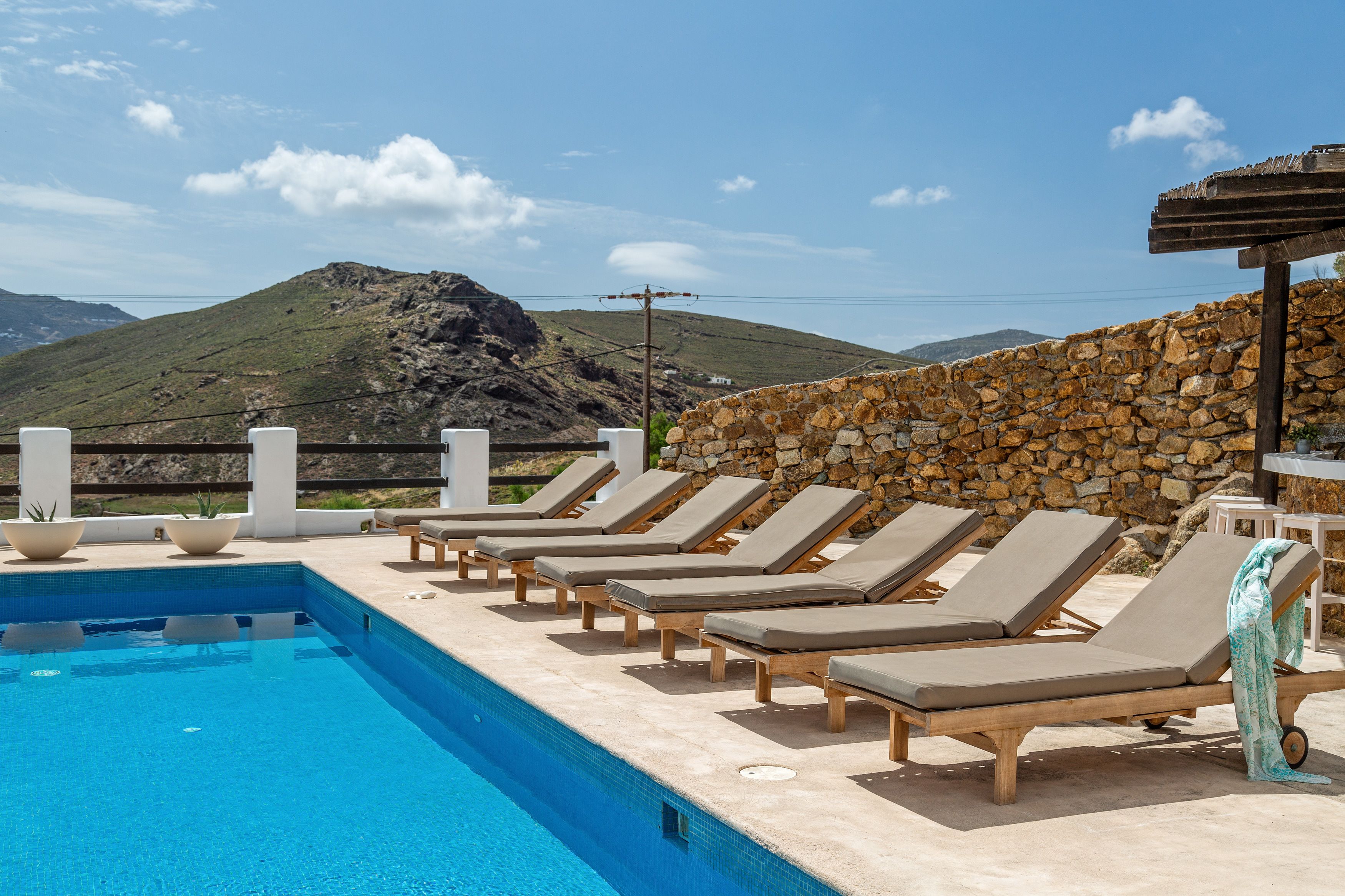 Zona junto a la piscina con sillones de descanso y un muro de piedra, con vistas a un paisaje montañoso.