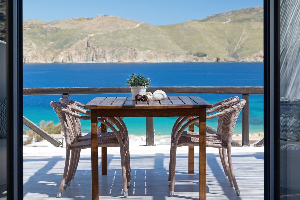 A wooden table with wicker chairs on a balcony overlooking a coastal view.