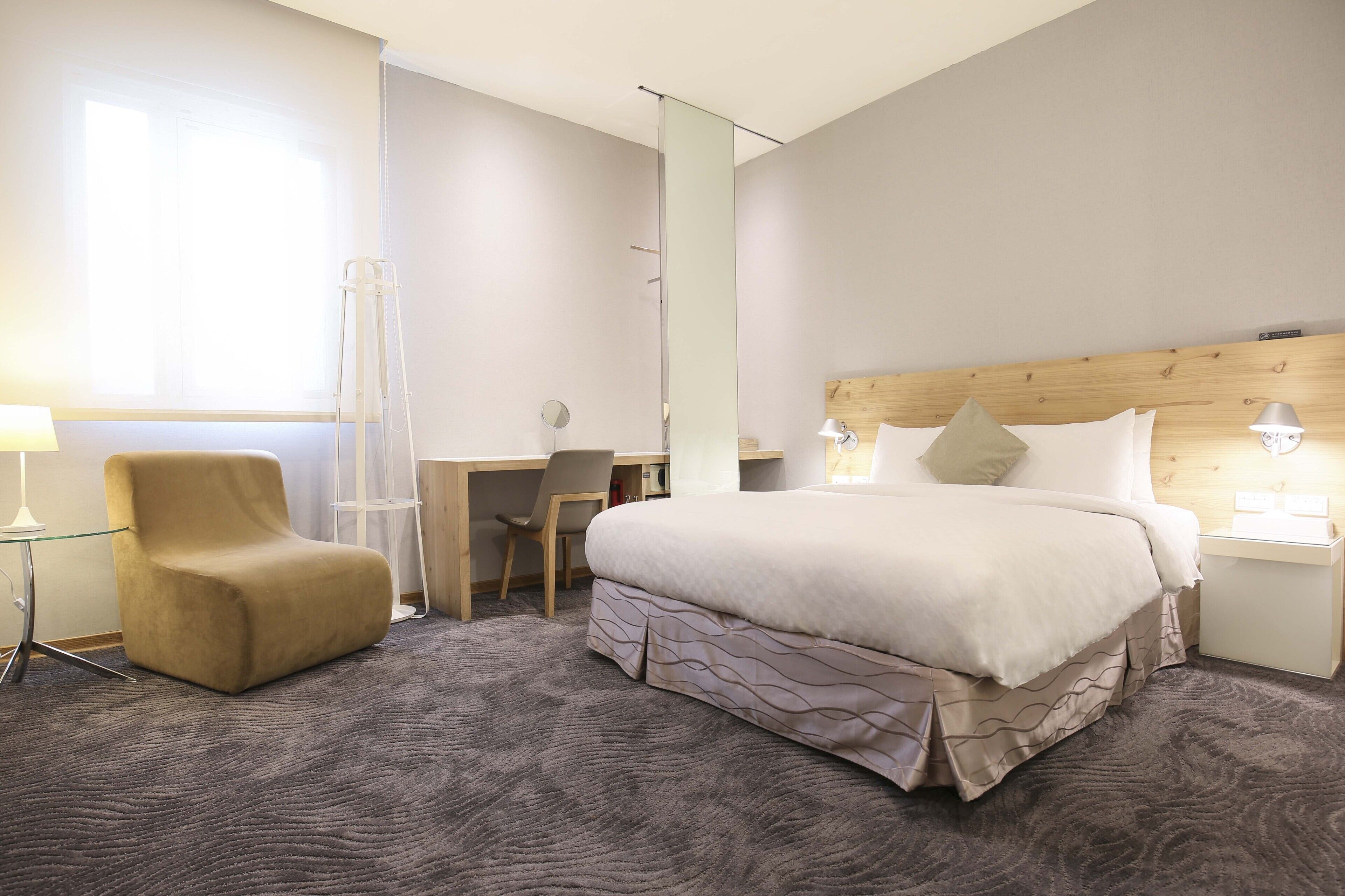 A hotel room with a large bed, a desk, a chair, and a standing lamp.