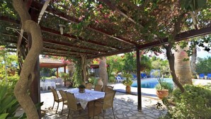 An outdoor patio with a stone floor, a table with chairs, and a pergola with climbing plants. - Follow The Sun (Polis)