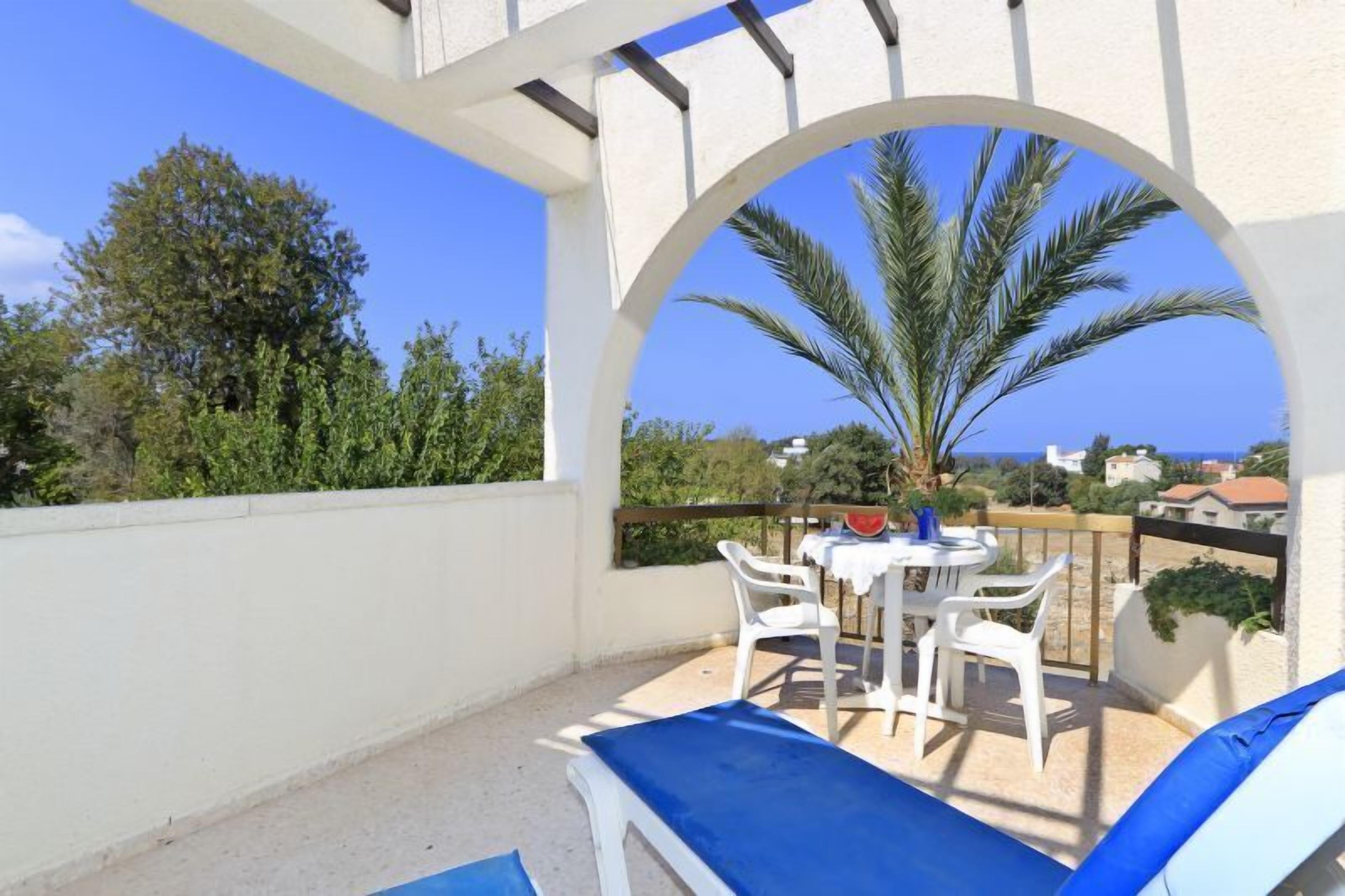 A balcony with a white table and chairs, a blue cushion, and a view of palm trees and houses.