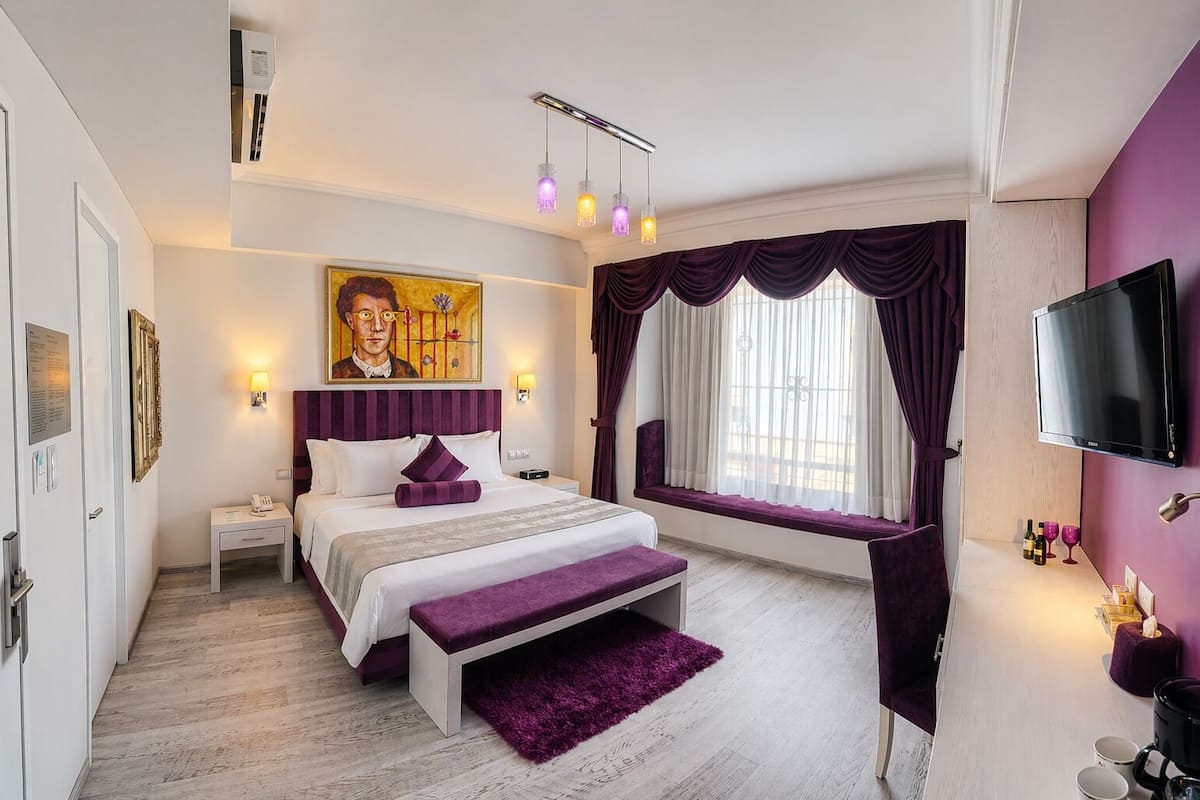 Un dormitorio con cama, mesita de noche, lámpara, una obra de arte, ventana con cortinas y televisor.