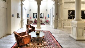 A spacious lobby with high ceilings, white columns, and a patterned rug. There are seating areas with chairs and a table, and artwork on the walls. - Hotel Andante (Puebla)