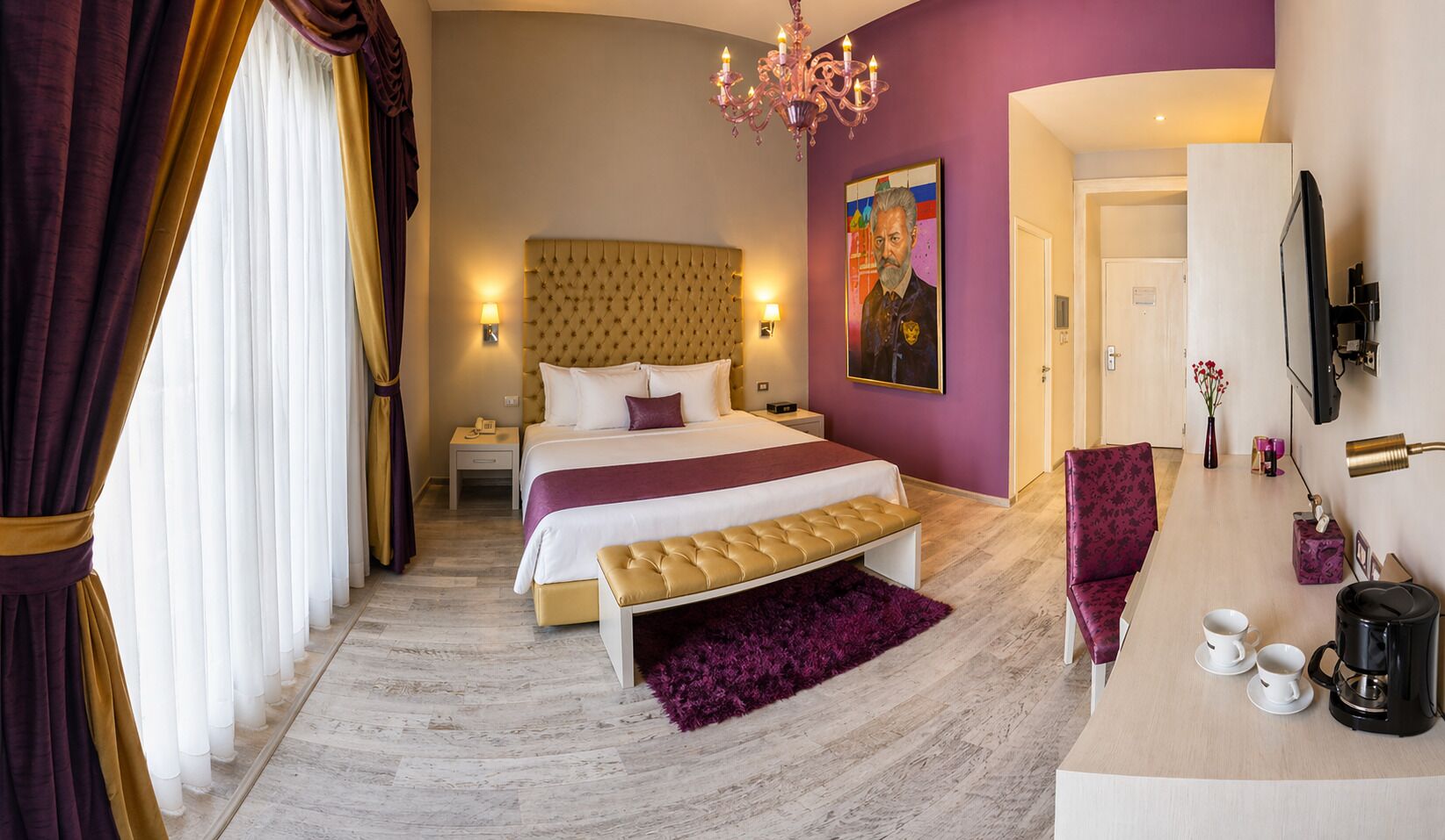 A bedroom with a large bed, a bench, a chair, a nightstand, a bedside table, a chandelier, a painting, a mirror, and a coffee maker.