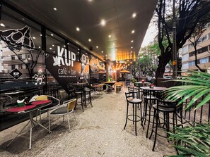 An outdoor café with tables and chairs, a large sign reading "KUP café + bar", and a cobblestone floor. - Royal Design Savassi (Belo Horizonte)