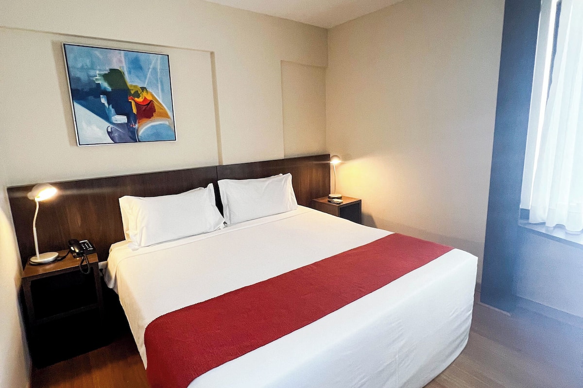 A hotel room with a large bed, a nightstand with a lamp, a painting on the wall, and a window with curtains.