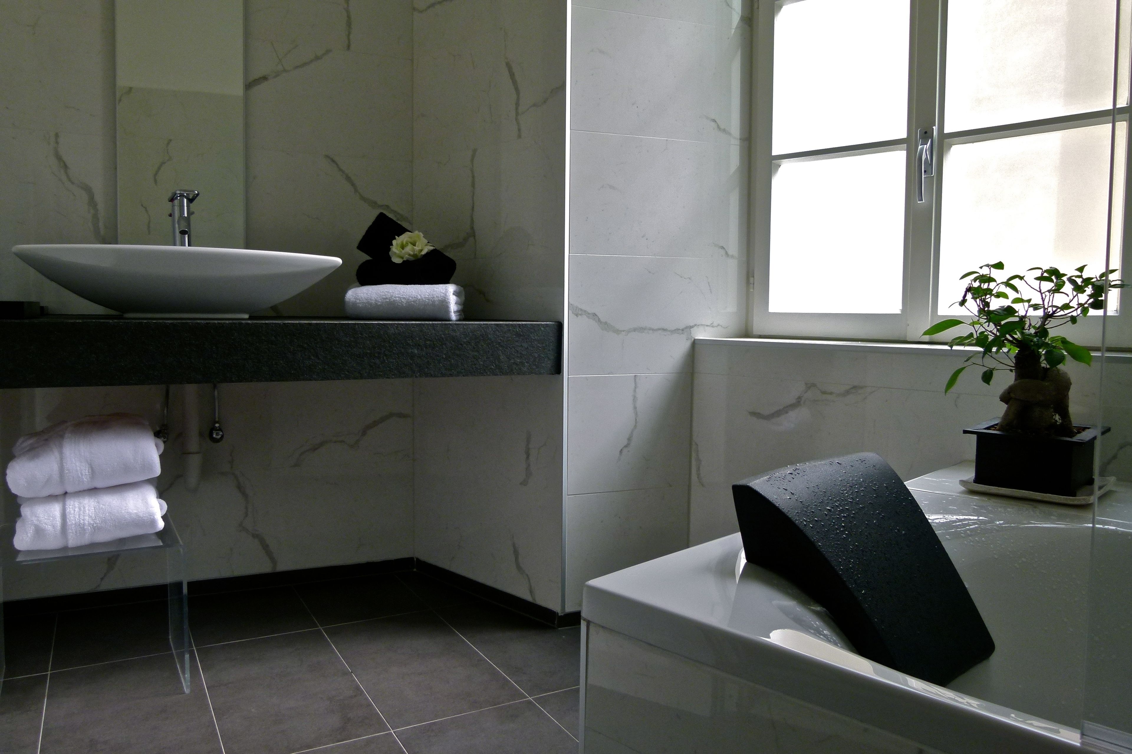 Une salle de bain avec un lavabo blanc, des murs en marbre, un porte-serviettes noir et une fenêtre laissant entrer la lumière naturelle.