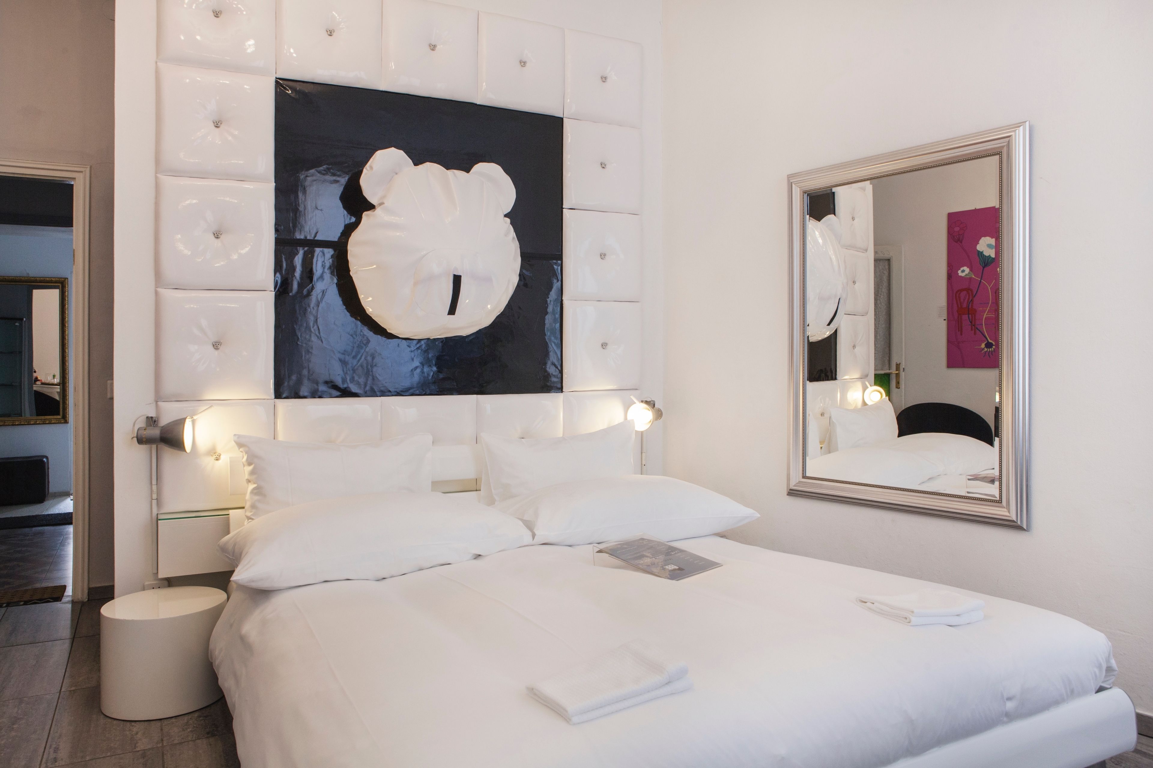 A modern hotel room with a large bed, a headboard with a bear design, a round bedside table, and a mirror on the wall.