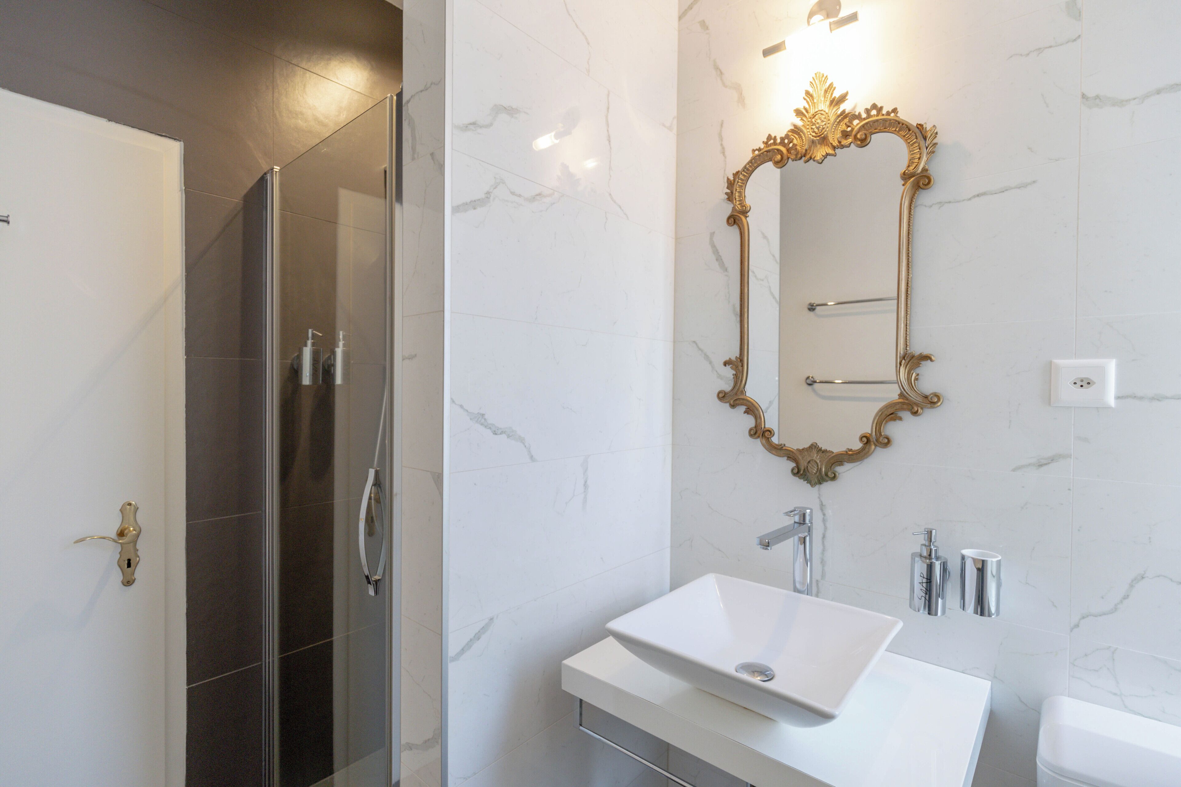 A modern bathroom with a glass-enclosed shower, a white sink, and a gold-framed mirror.