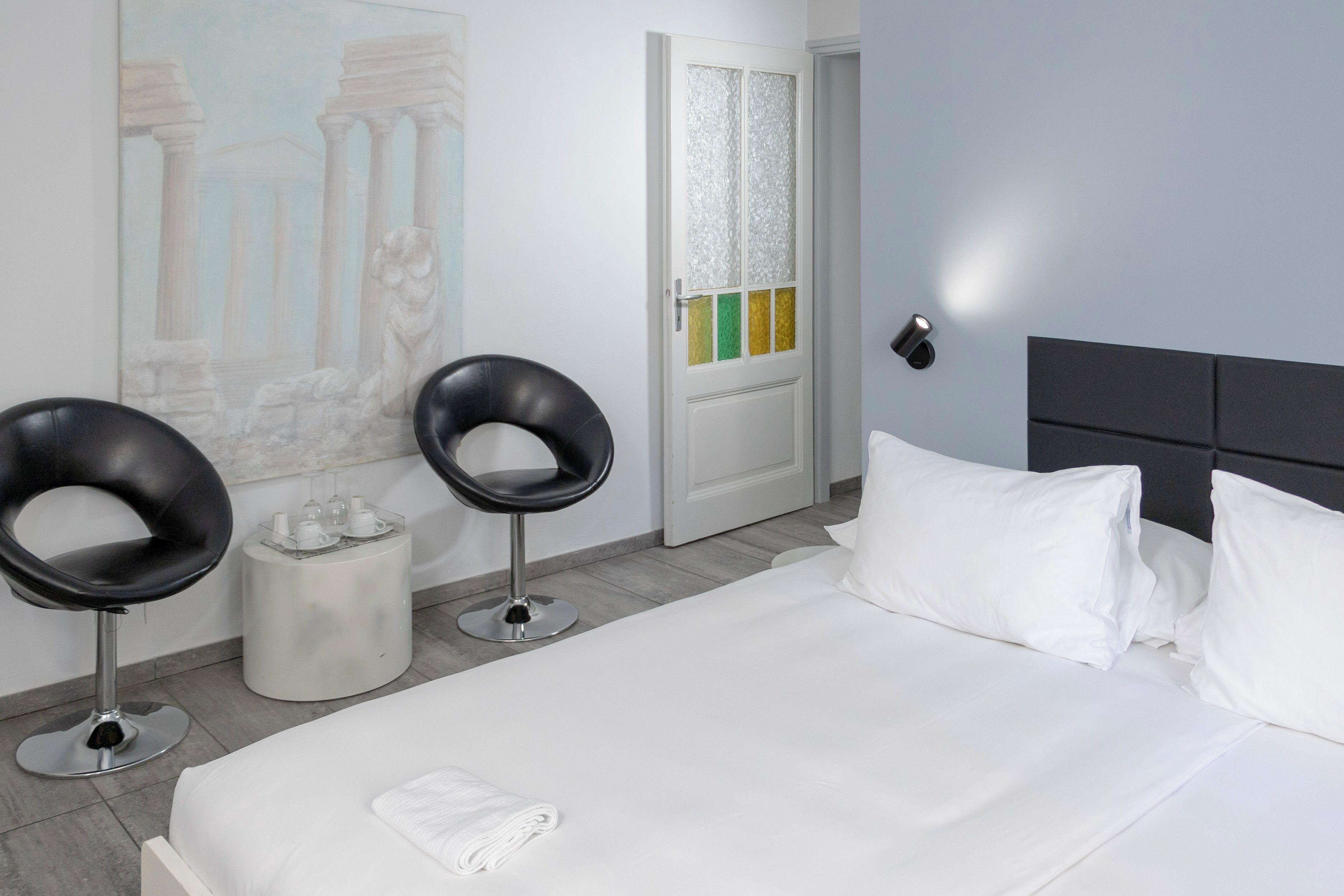 Une chambre d’hôtel avec un lit, deux chaises noires, un tableau, une porte à panneaux colorés et une table de chevet.