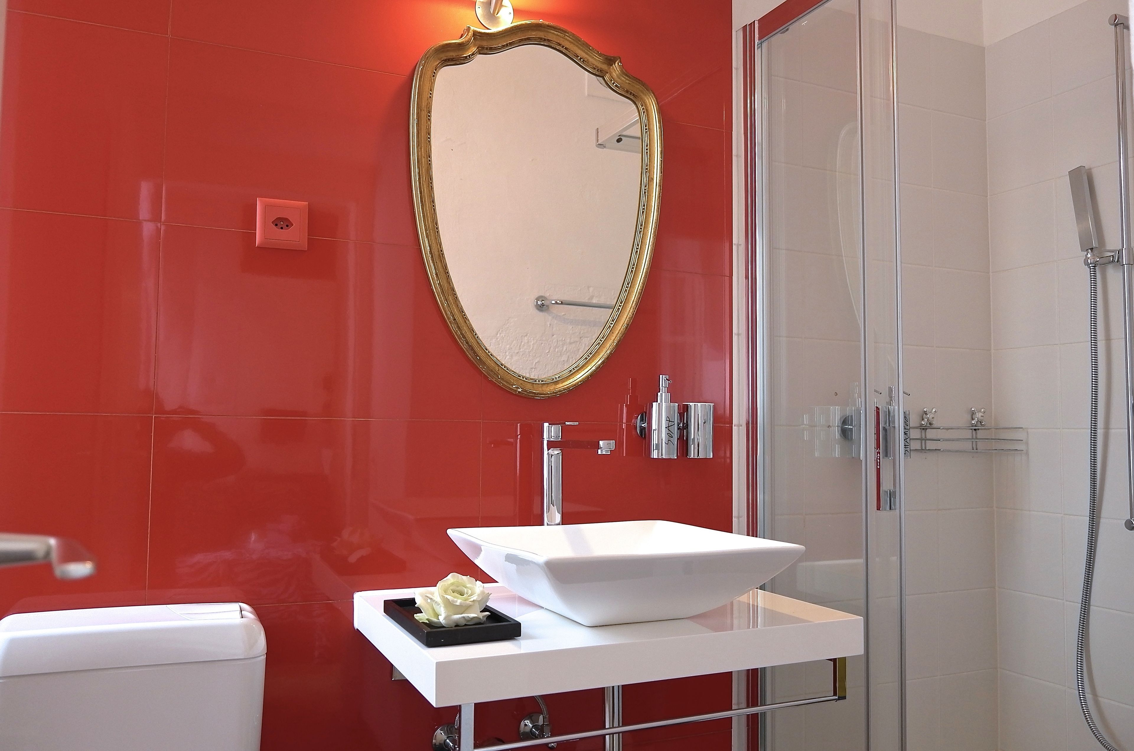 Un baño con un lavamanos blanco, un espejo con marco dorado y una pared con azulejos rojos.