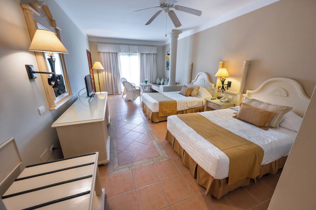 A hotel room with two beds, a desk, a chair, a ceiling fan, and a window with curtains.