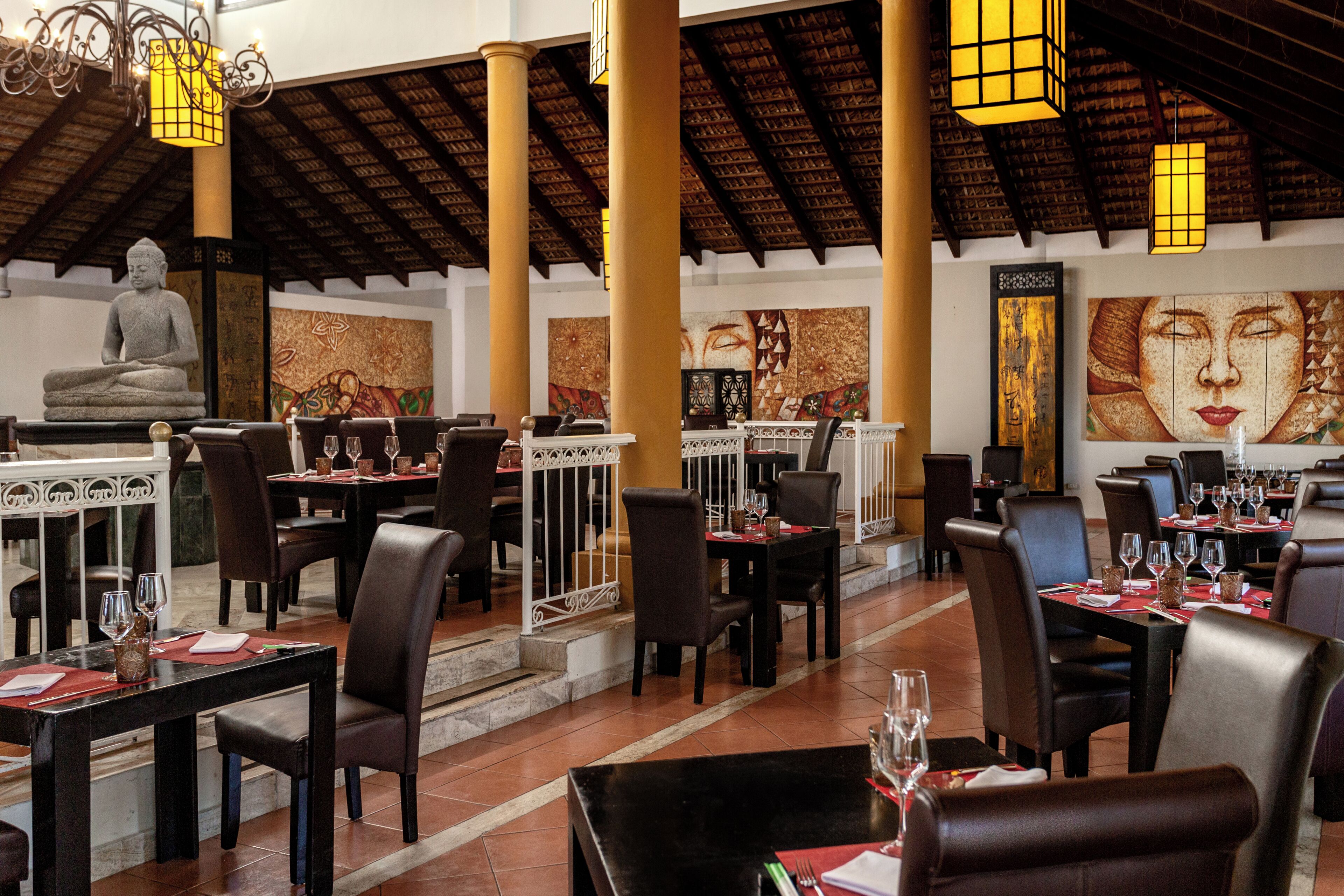 A restaurant with tables set for dining, a Buddha statue, and decorative wall art.